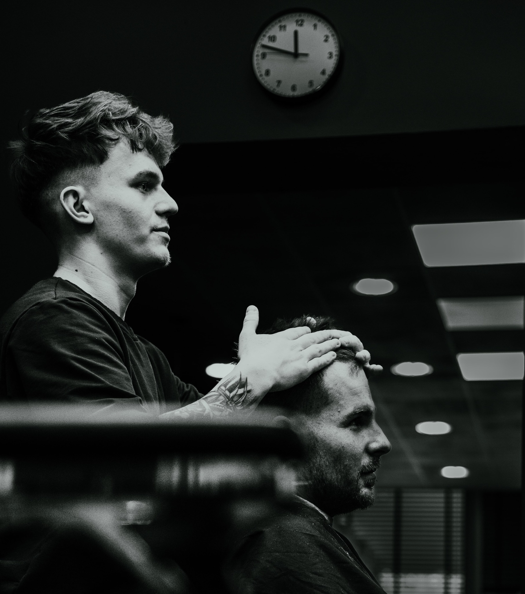 Fryzjer stylizuje włosy klienta w salonie, widoczny zegar na ścianie i tatuaż na ramieniu stylisty.