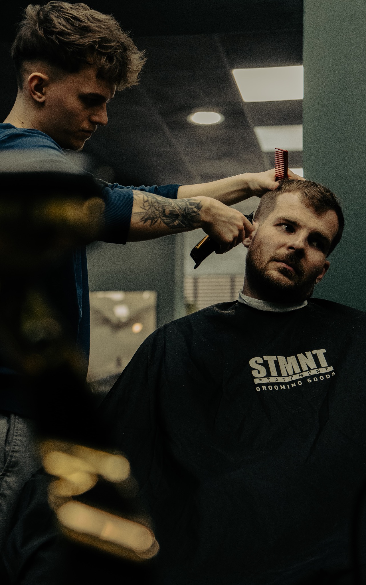 Fryzjer z tatuażem na ramieniu strzyże klienta maszynką, klient ma na sobie pelerynę z napisem STMN Grooming Goods.