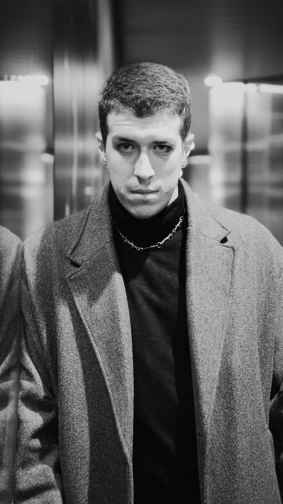 Portret młodego mężczyzny w szarym płaszczu i czarnym golfie, z łańcuszkiem na szyi, na tle rozmytego wnętrza.