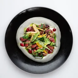 Kompozycja dania na czarnym talerzu: gnocchi z buraków, kremowy sos, rukola, pestki dyni i marynowane warzywa, oświetlone od góry.