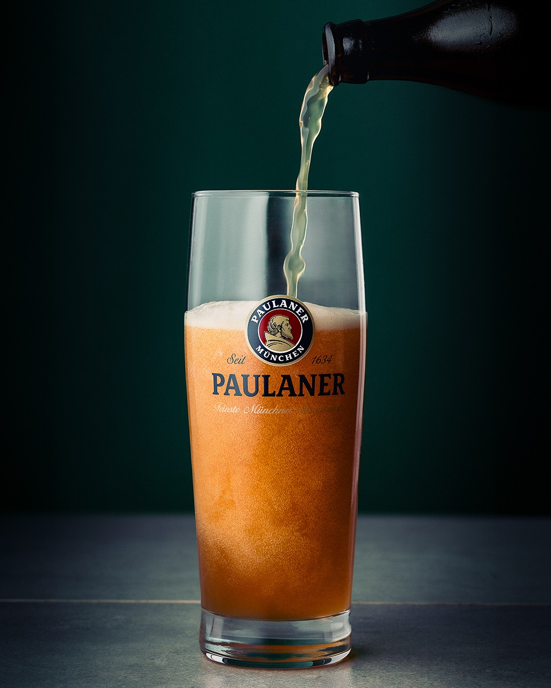Nalewanie piwa pszenicznego Paulaner do szklanki, widoczny logo i charakterystyczna mętność trunku.