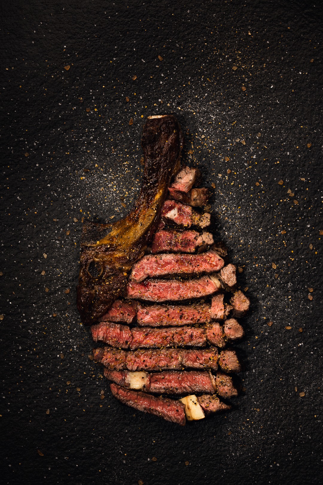 Krojony stek Tomahawk z kością, ułożony na czarnym kamiennym tle posypanym solą i przyprawami.