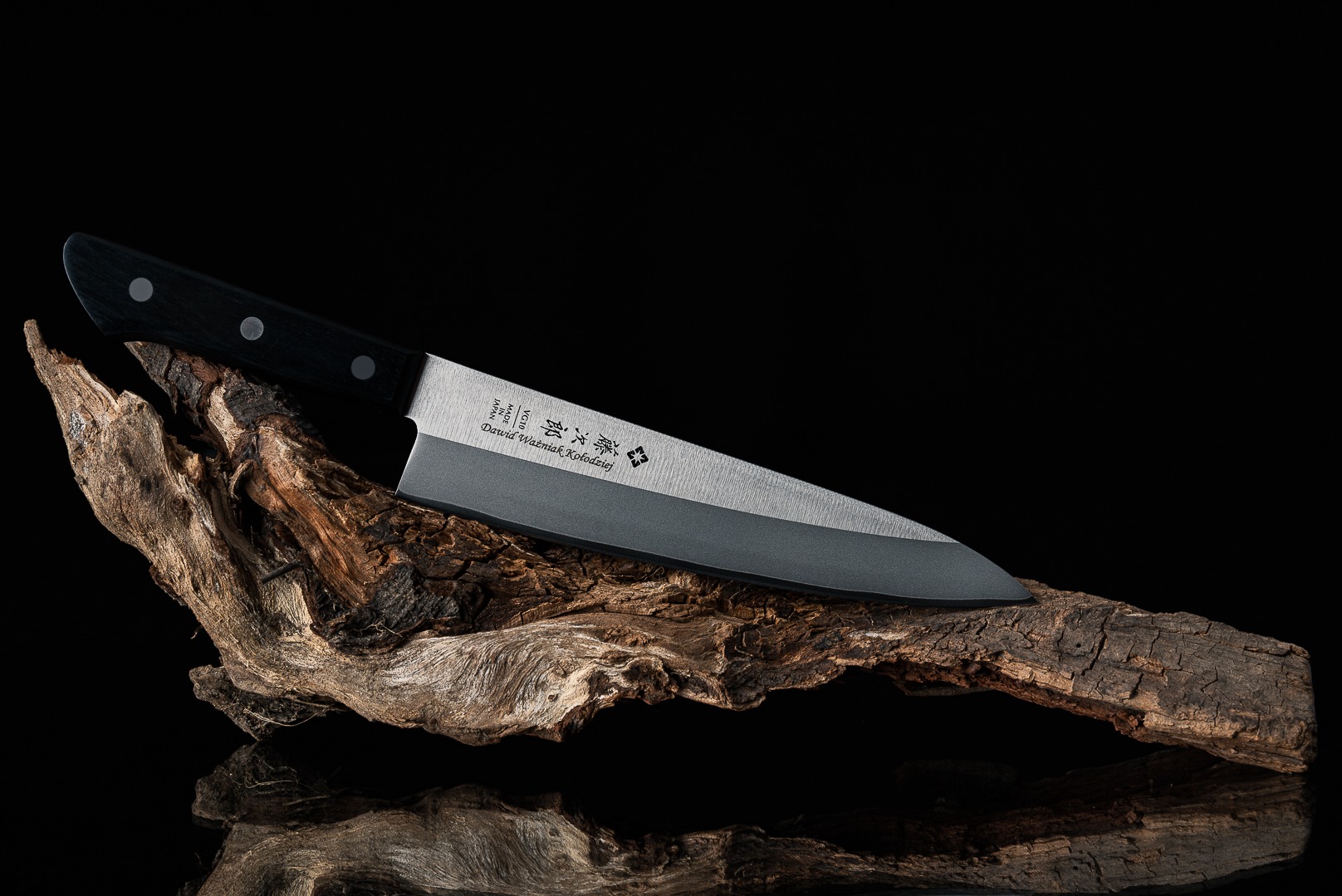 Ostrze japońskiego noża kuchennego leży na kawałku starego drewna na czarnym tle z odbiciem.