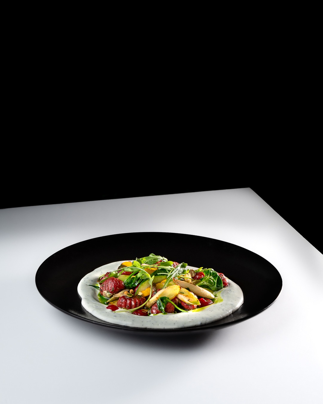 Apetyczna sałatka z pieczonych buraków, gruszek, rukoli i orzechów piniowych, podana na czarnym talerzu z sosem jogurtowym na białym tle.