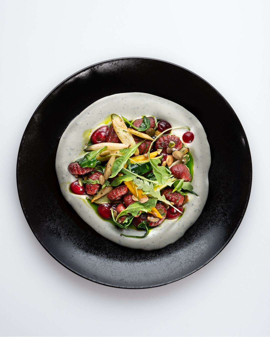 Kompozycja dania na czarnym talerzu: gnocchi z buraków, kremowy sos, rukola, pestki dyni i marynowane warzywa, oświetlone od góry.