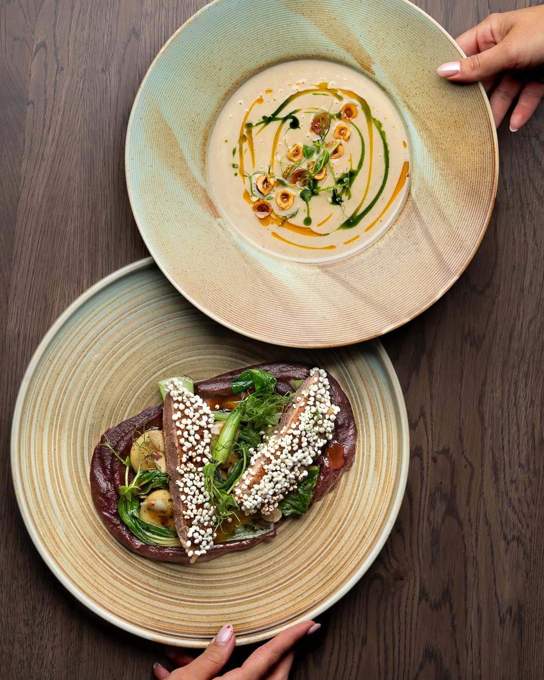 Dwa talerze z daniami na drewnianym stole: kremowa zupa z orzechami i ziołami oraz mięso z warzywami i białymi kuleczkami, trzymane przez dłonie z delikatnym różowym manicure.