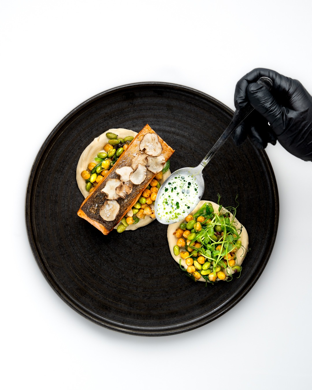 Danie z łososiem, ciecierzycą, edamame, grzybami i sosem, serwowane na czarnym talerzu, dekorowane przez kucharza w czarnych rękawiczkach.