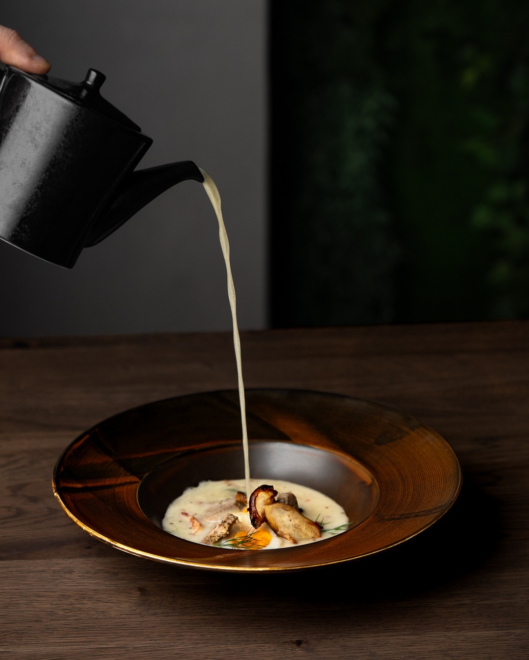 Eleganckie ujęcie zupy krem z grzybami, polewanej z czarnego dzbanka, na drewnianym stole w Krakowie. Złota obwódka talerza i minimalistyczne tło.
