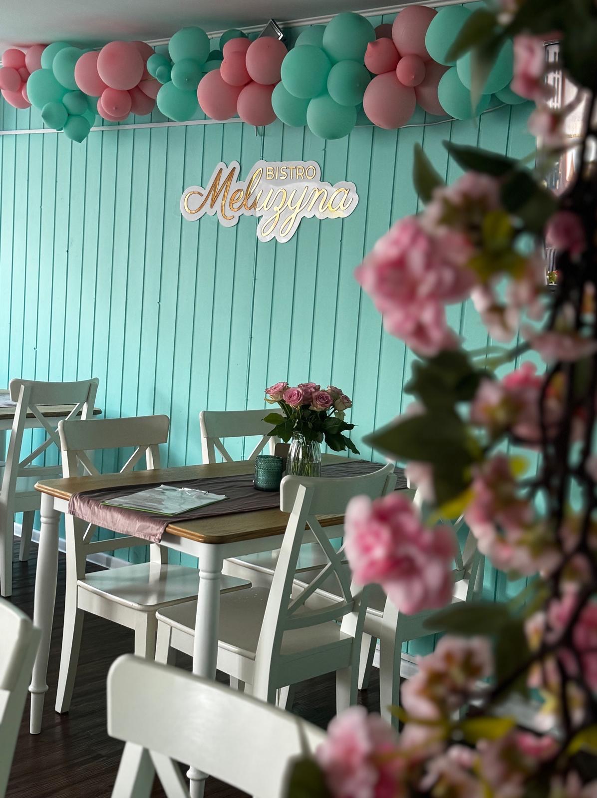 Uroczysty wystrój wnętrza bistro z turkusowymi ścianami, balonami w kolorach różu i mięty, stołami z białymi krzesłami, bukietem róż w wazonie i dekoracją kwiatową z przodu