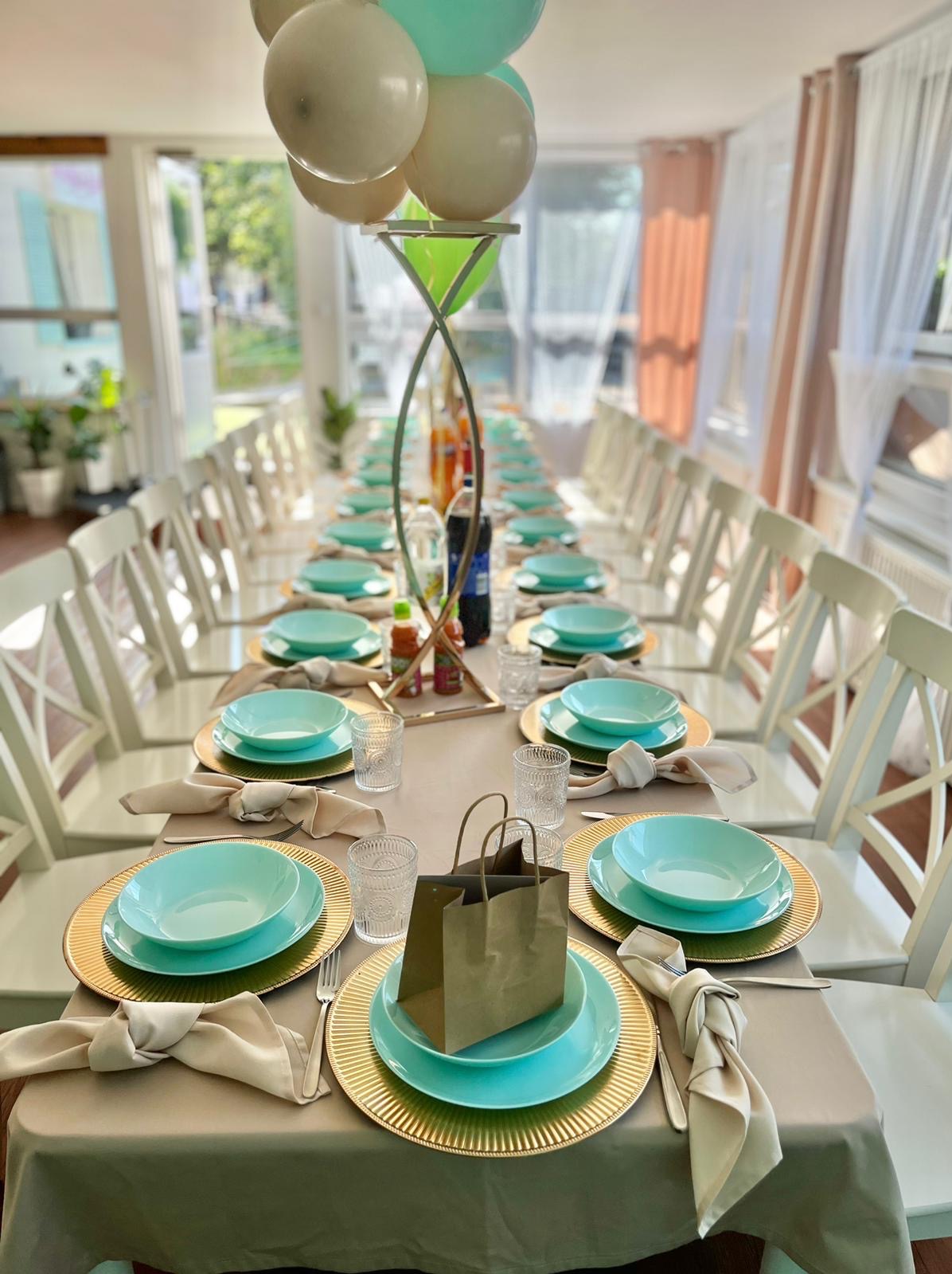 Długi stół nakryty beżowym obrusem, zastawiony złotymi podkładkami, turkusowymi talerzami, szklankami i beżowymi serwetkami. Na każdym nakryciu stoi brązowa torebka upominkowa. Nad stołem unoszą...