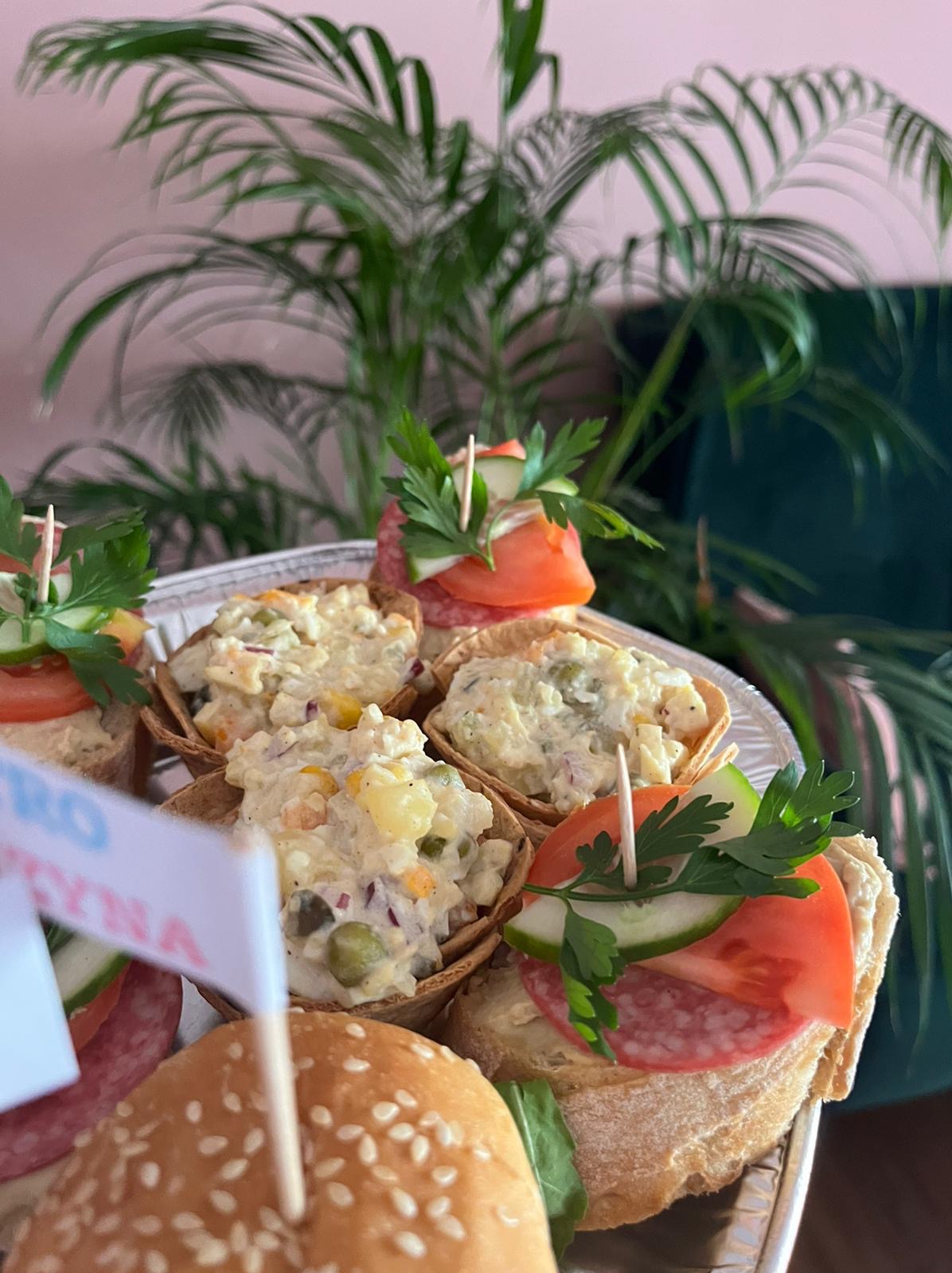 Taca z przekąskami: mini tartaletki z sałatką jarzynową, kanapki z salami, pomidorem, ogórkiem i pietruszką, oraz mini burgery, na tle zielonej rośliny doniczkowej.