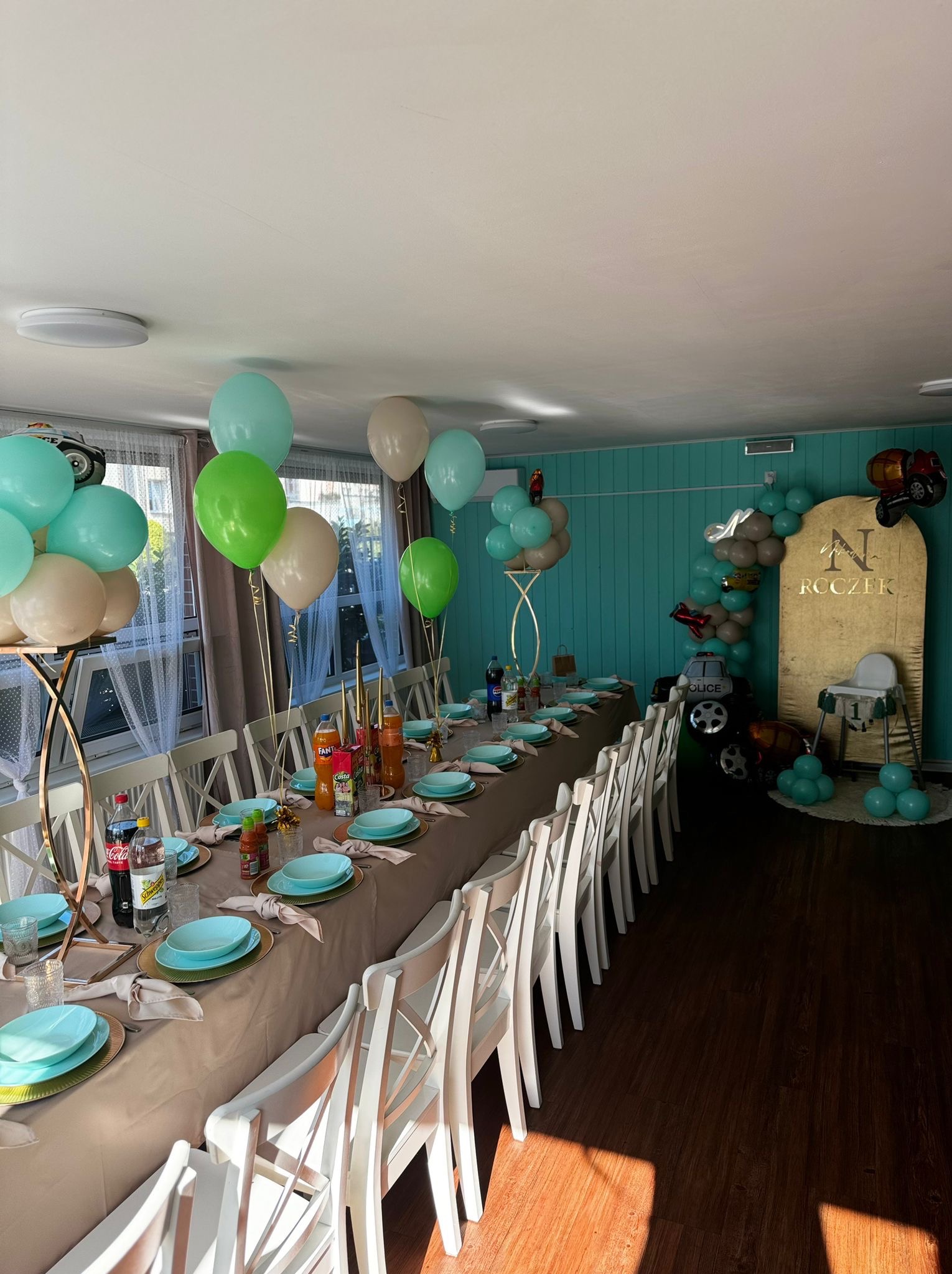 Uroczysty stół z turkusową zastawą i dekoracjami z balonów w odcieniach zieleni i beżu, przygotowany na przyjęcie urodzinowe dziecka z motywem samochodów.