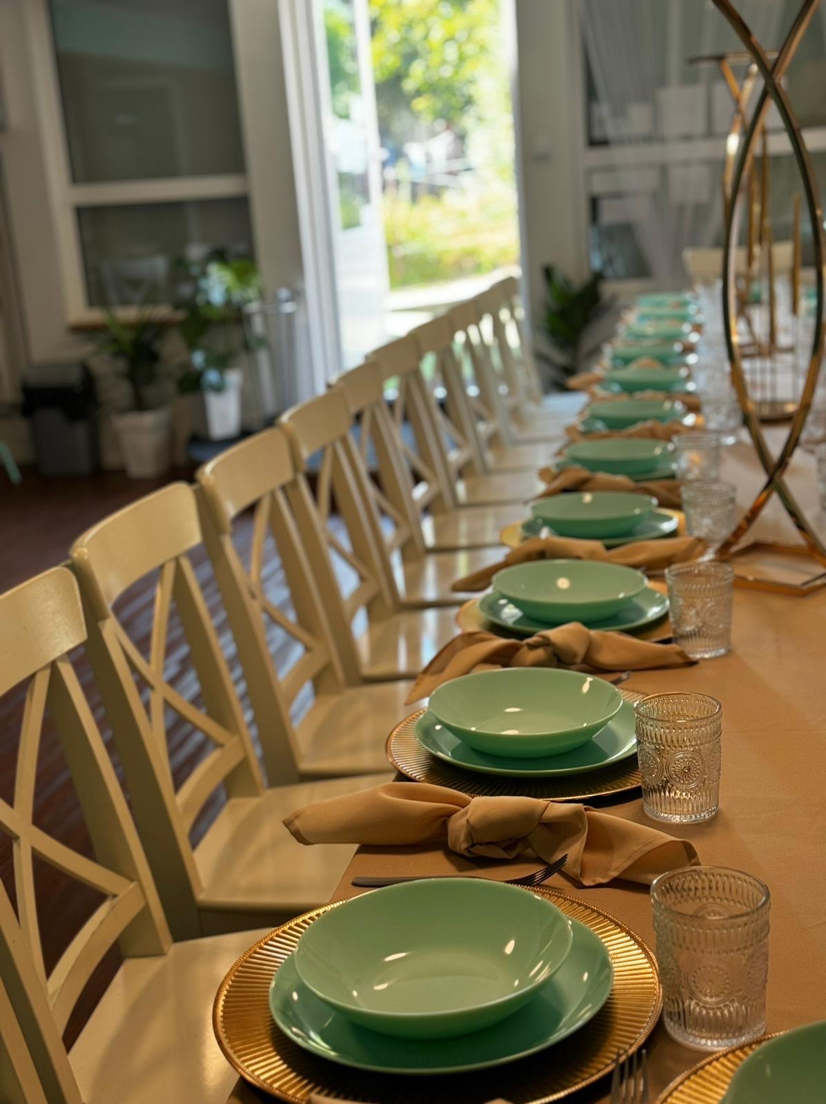 Długi stół z beżowym obrusem, nakryty dla wielu gości: złote podkładki, turkusowe talerze i miski, beżowe serwetki, szklanki z tłoczonym wzorem, białe krzesła z krzyżowym oparciem, wysokie metalowe...