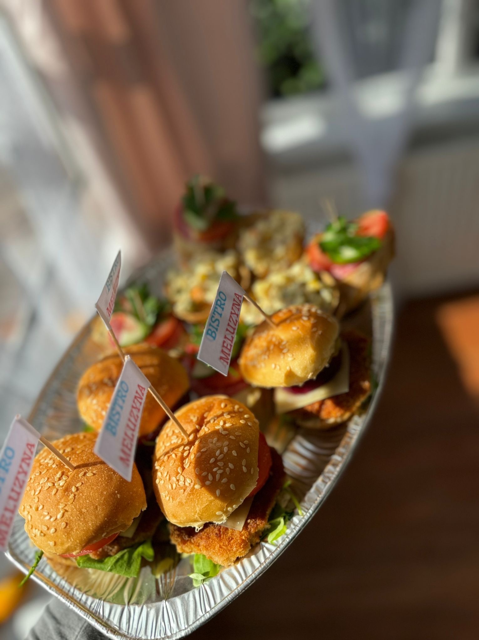 Srebrna tacka z mini burgerami i kanapeczkami, każdy burger oznaczony małą flagą z napisem 'Bistro Meluzyna', oświetlone naturalnym światłem z okna.