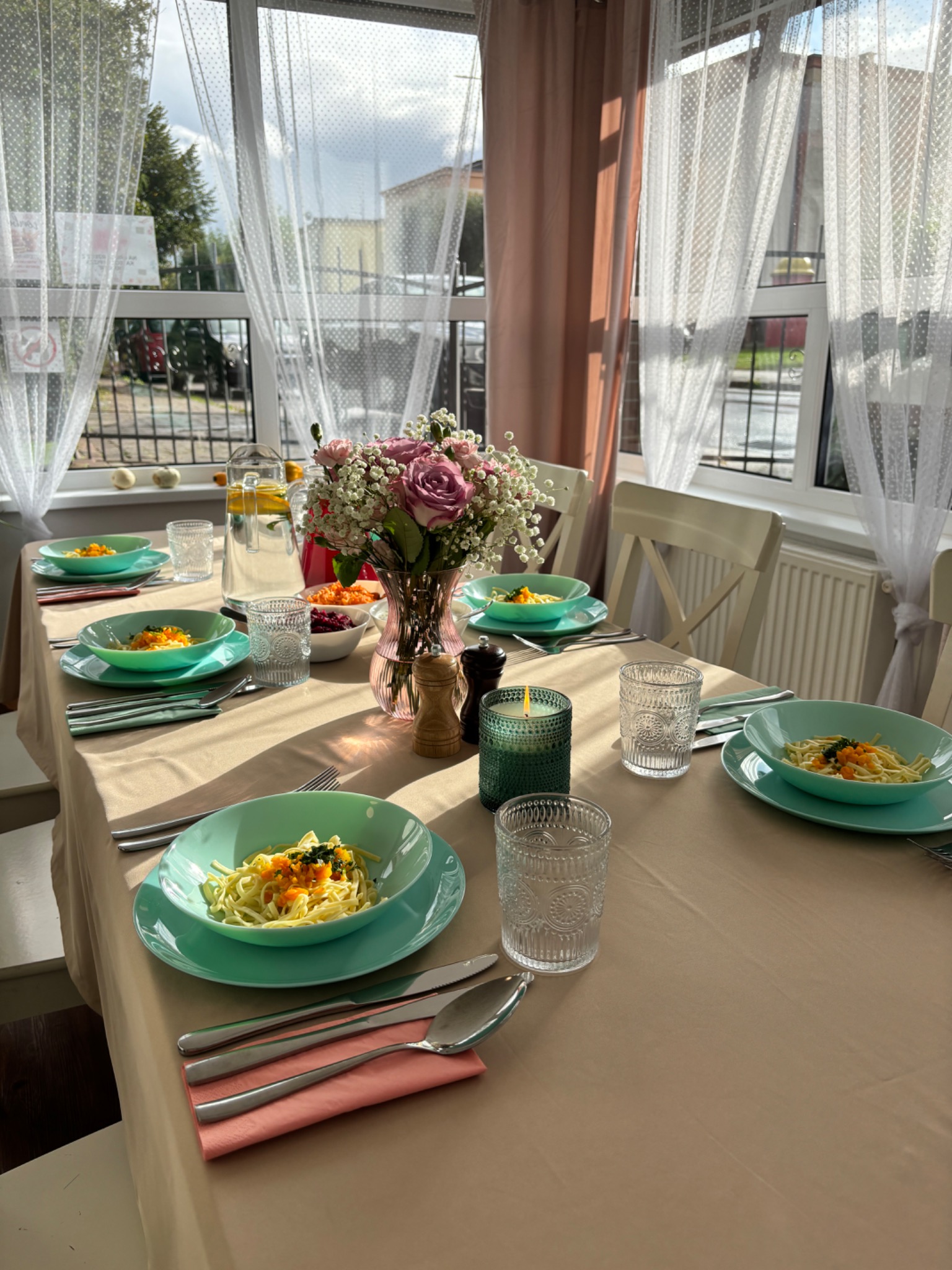 Uroczysty stół nakryty beżowym obrusem, z turkusową zastawą, daniem makaronowym, szklankami z tłoczonym wzorem, bukietem różowych róż w wazonie, świecą w szklanym świeczniku i dzbankiem z wodą...