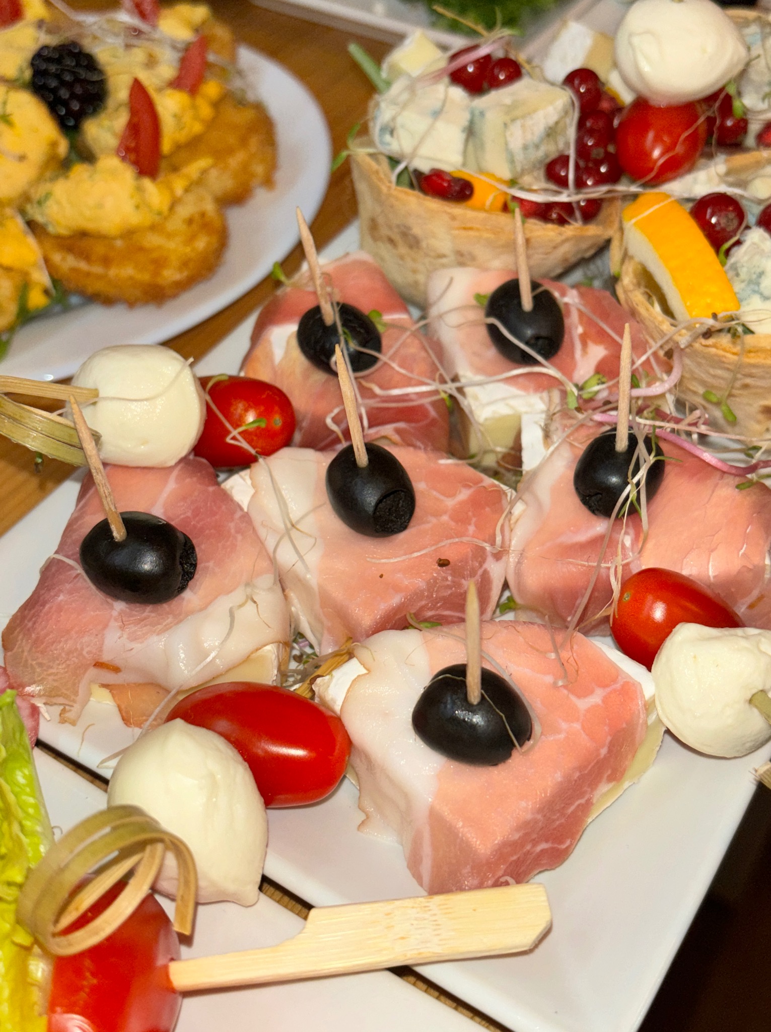 Zbliżenie na talerz z różnorodnymi przekąskami: szynka parmeńska z oliwkami i mini-mozzarellą na wykałaczkach, obok sałatka z serem pleśniowym i czerwonymi porzeczkami w jadalnych koszyczkach, a...