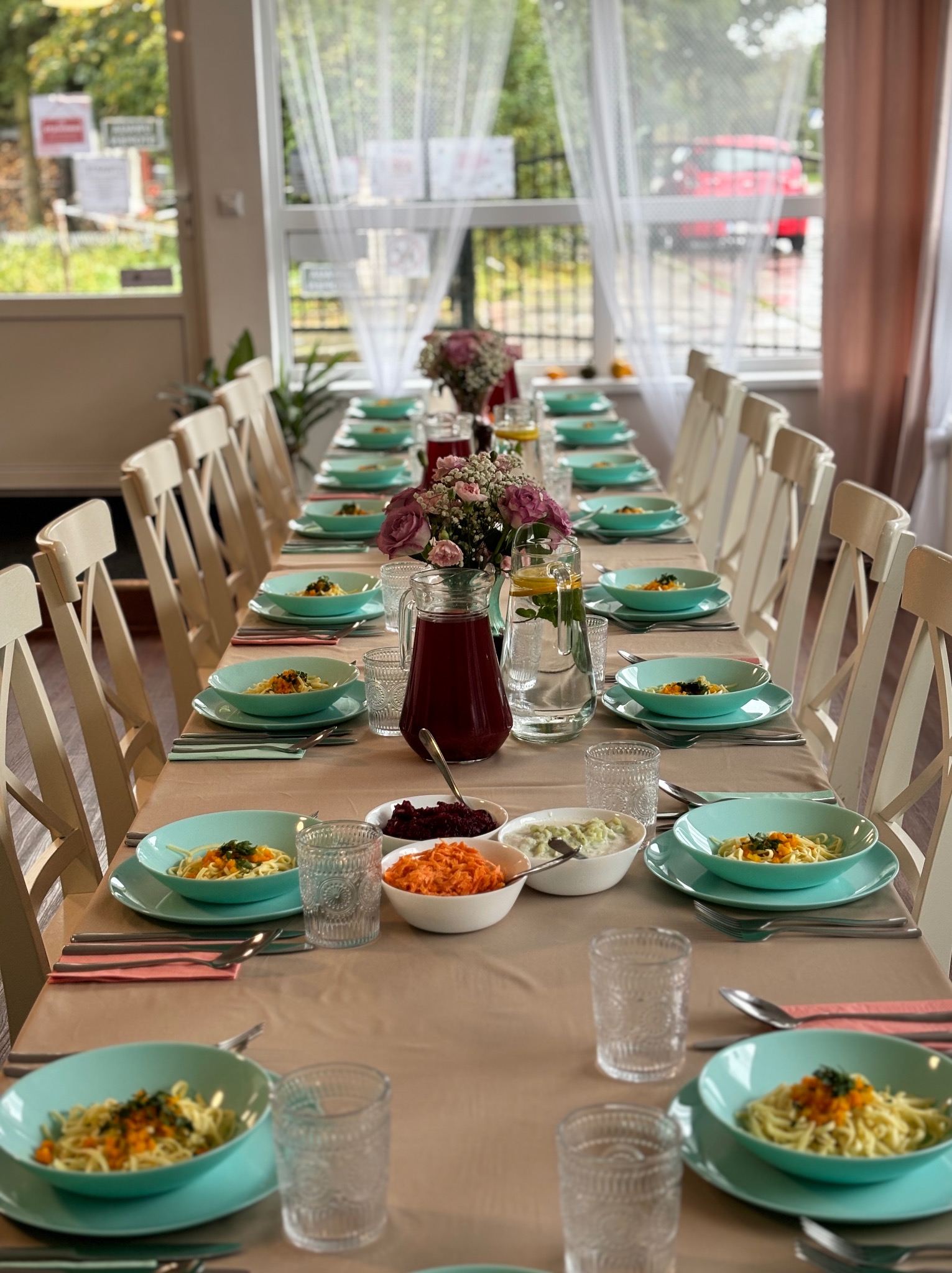 Długi stół nakryty na przyjęcie, z turkusową zastawą, na którym znajdują się dania z makaronem, sałatki i napoje w dzbankach, ustawiony w jasnym pomieszczeniu z oknami.