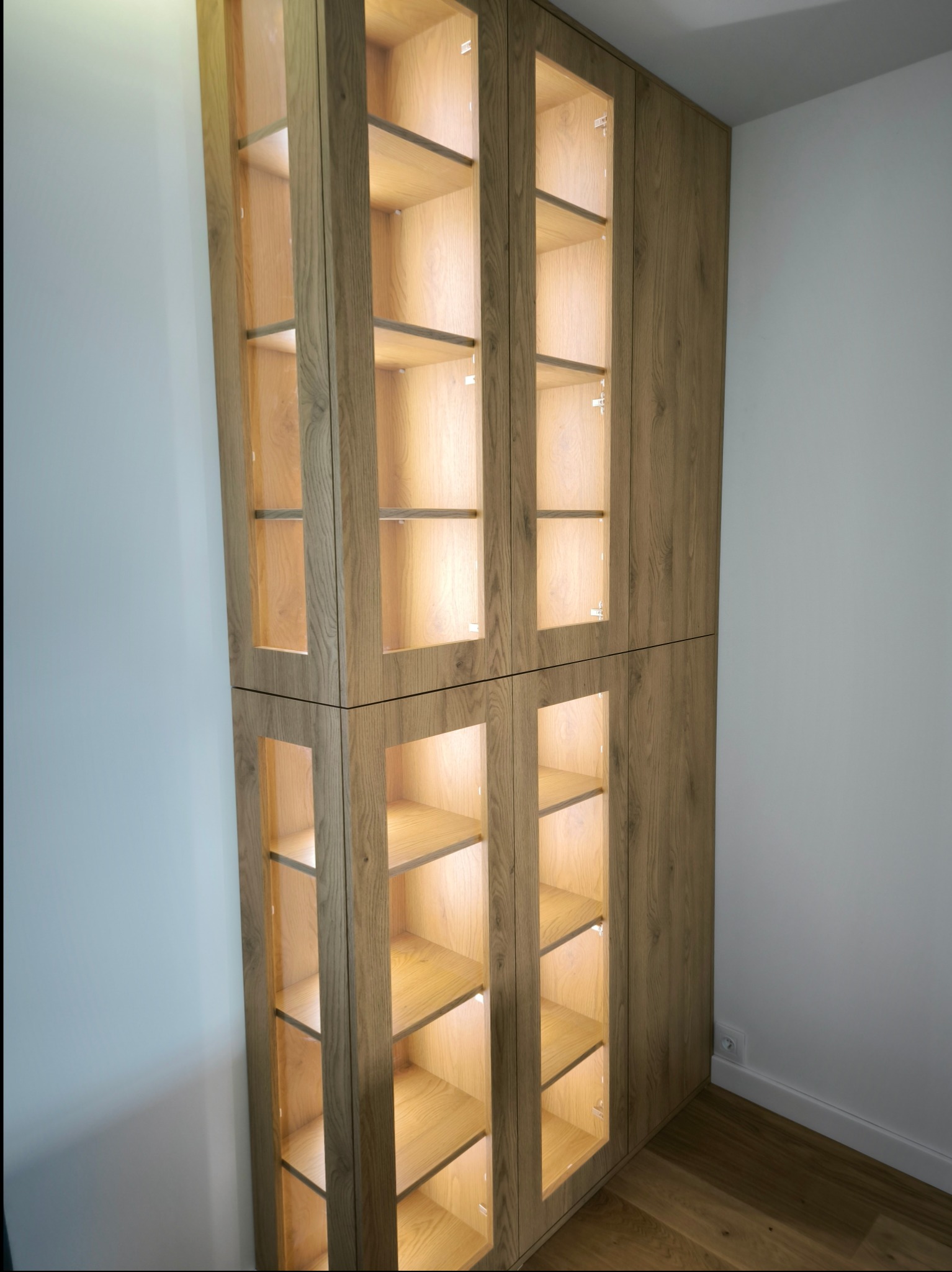 Wysoka, dębowa szafa z przeszklonymi drzwiczkami i oświetleniem LED wewnątrz, widoczna na tle białej ściany i drewnianej podłogi.