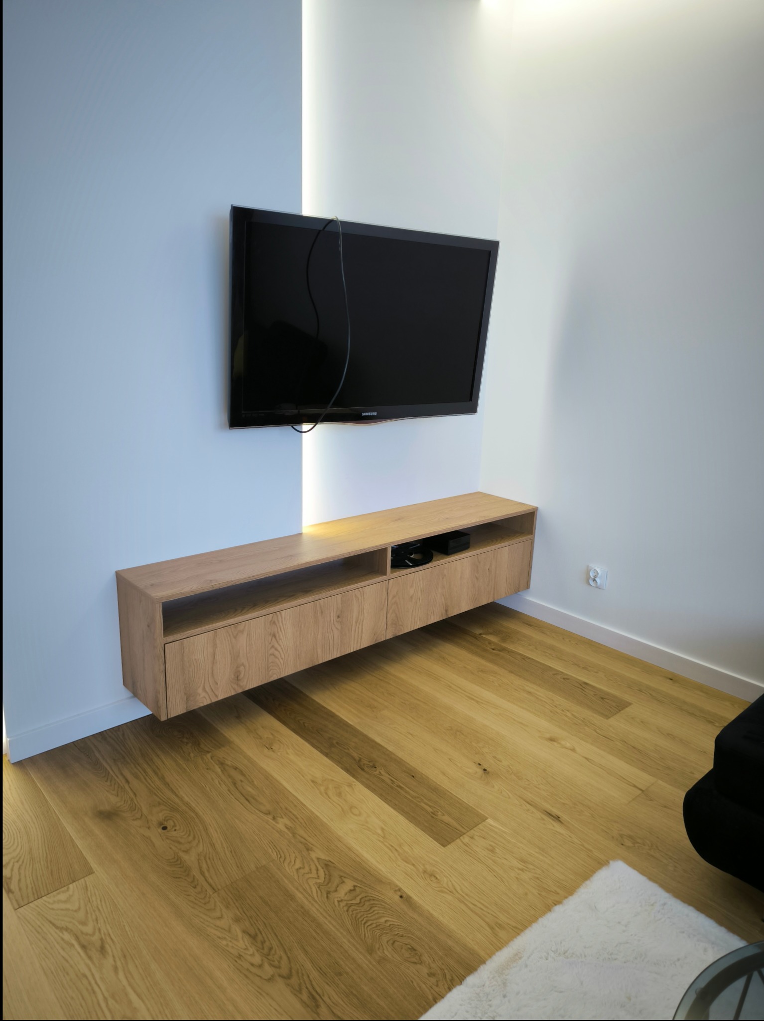 Minimalistyczna szafka RTV z jasnego drewna dębowego, zawieszona pod telewizorem na białej ścianie z wbudowanym oświetleniem LED, na tle drewnianej podłogi.