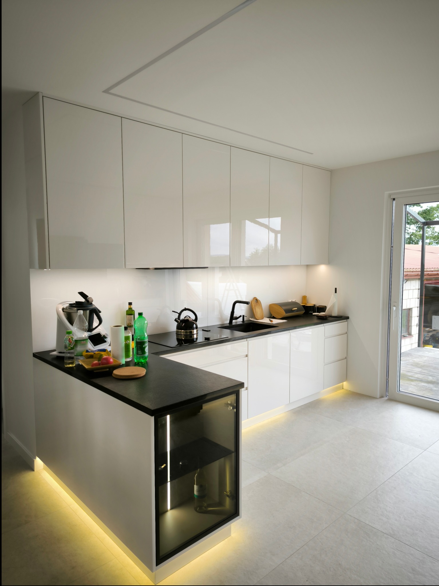Nowoczesna kuchnia z białymi, lakierowanymi frontami, czarnym blatem i oświetleniem LED pod szafkami, widok na przeszkloną szafkę z butelkami wina.