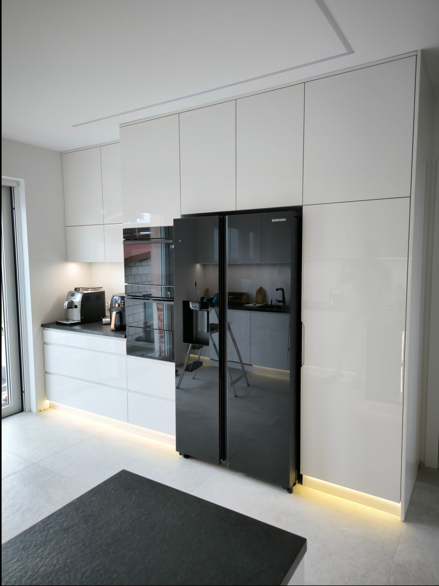 Biała, nowoczesna kuchnia z zabudową szafek pod sufit, czarnym blatem i lodówką Samsung side-by-side. Oświetlenie LED pod szafkami dolnymi. W odbiciu lodówki widoczny fragment wnętrza kuchni...