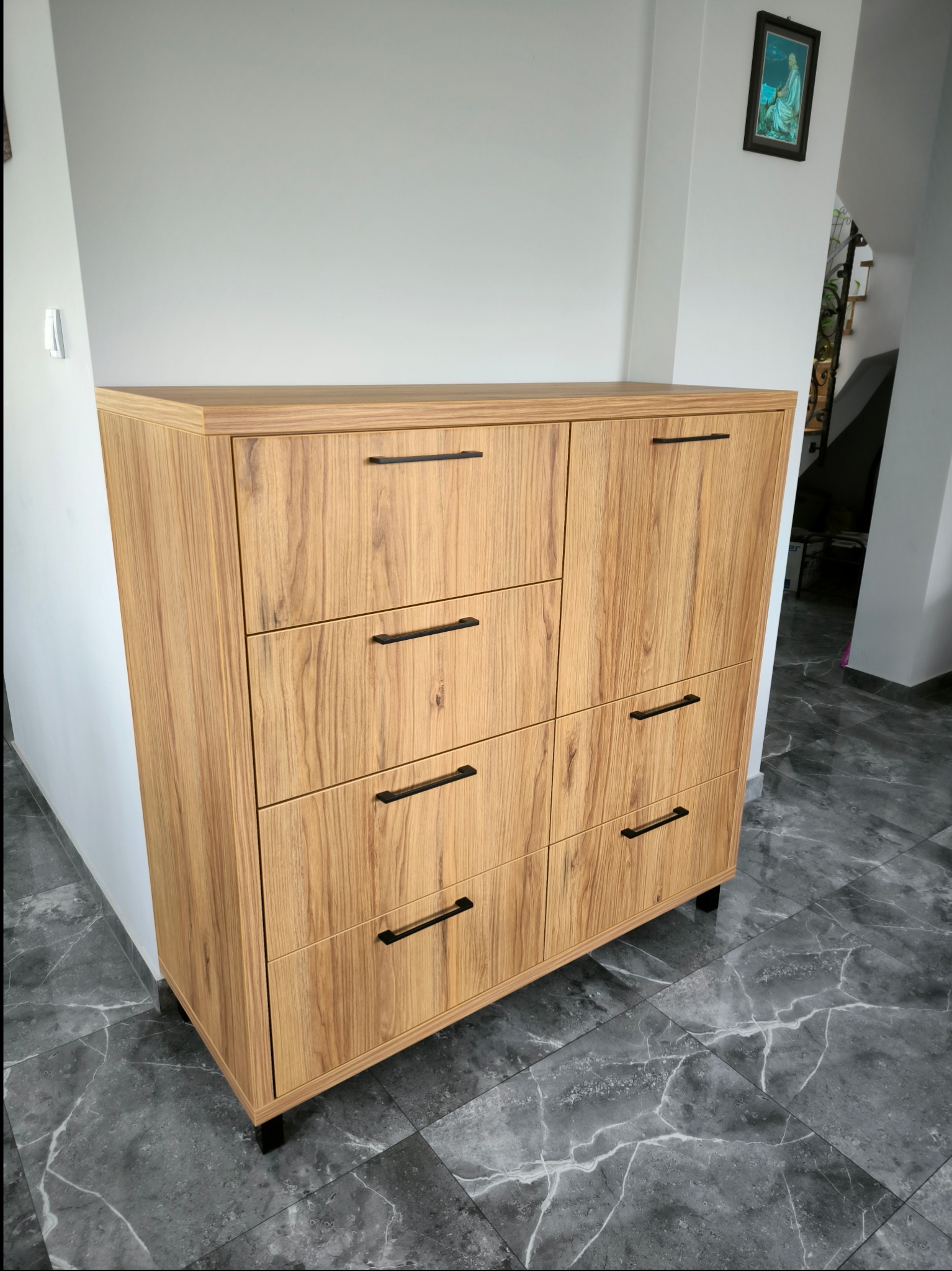 Drewniana komoda z czarnymi uchwytami na tle szarej marmurowej podłogi i białych ścian, z obrazem wiszącym w rogu.