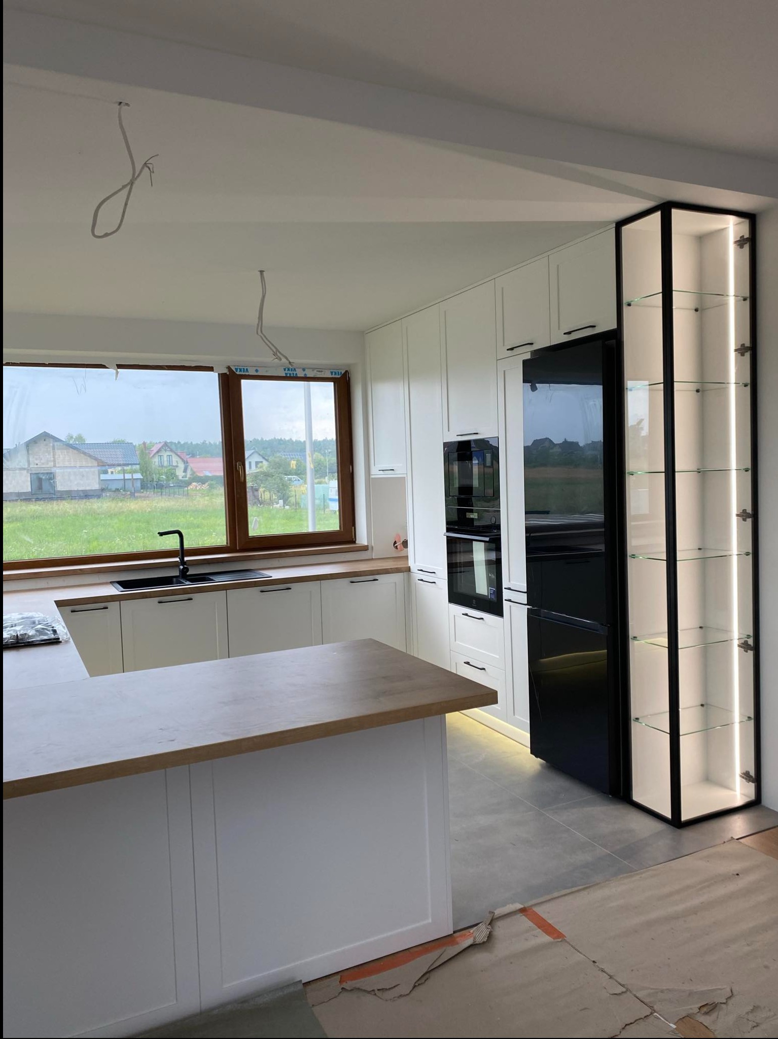 Nowoczesna, biała kuchnia z wyspą i czarnym sprzętem AGD, szklana witryna z podświetleniem LED, widoczne niedokończone prace wykończeniowe podłogi.