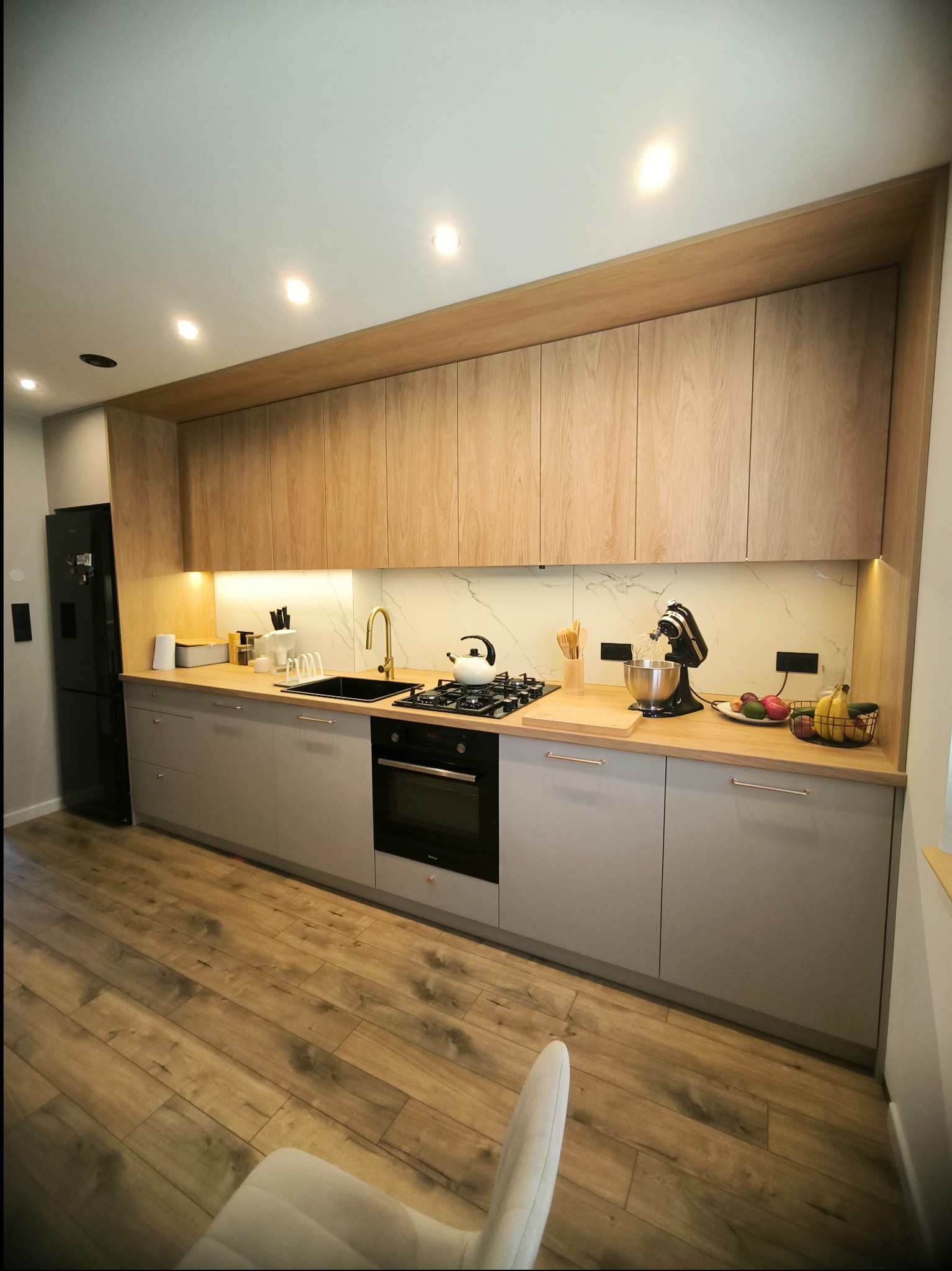 Nowoczesna kuchnia z drewnianymi górnymi szafkami i szarymi dolnymi, złota bateria, czarny zlew i sprzęt AGD, marmurowy backsplash, drewniana podłoga i fragment krzesła w rogu