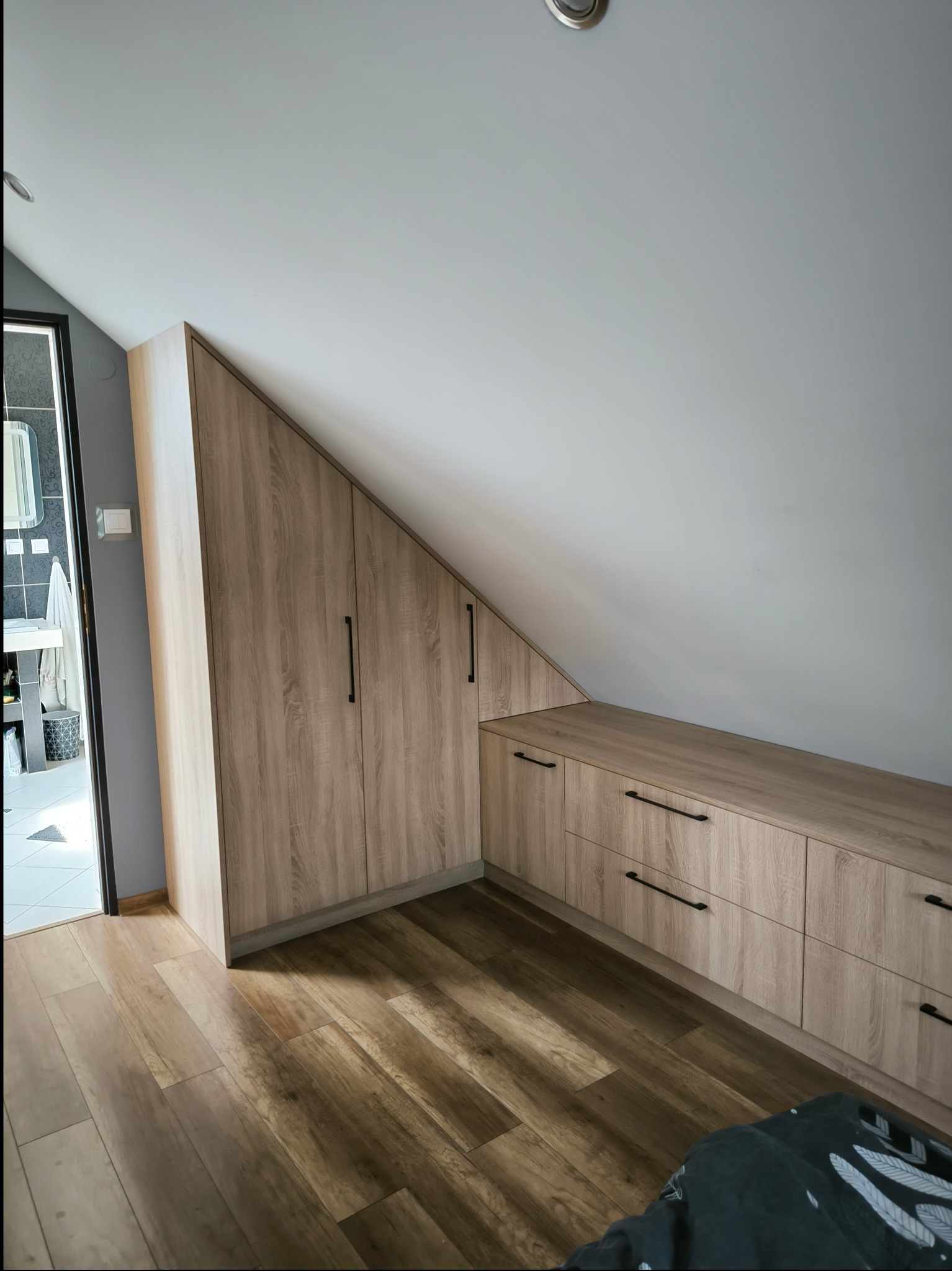 Jasna szafa wnękowa z drewnopodobnymi frontami i czarnymi uchwytami, zabudowa pod skosem na poddaszu, widoczna podłoga z paneli i fragment łazienki w tle.