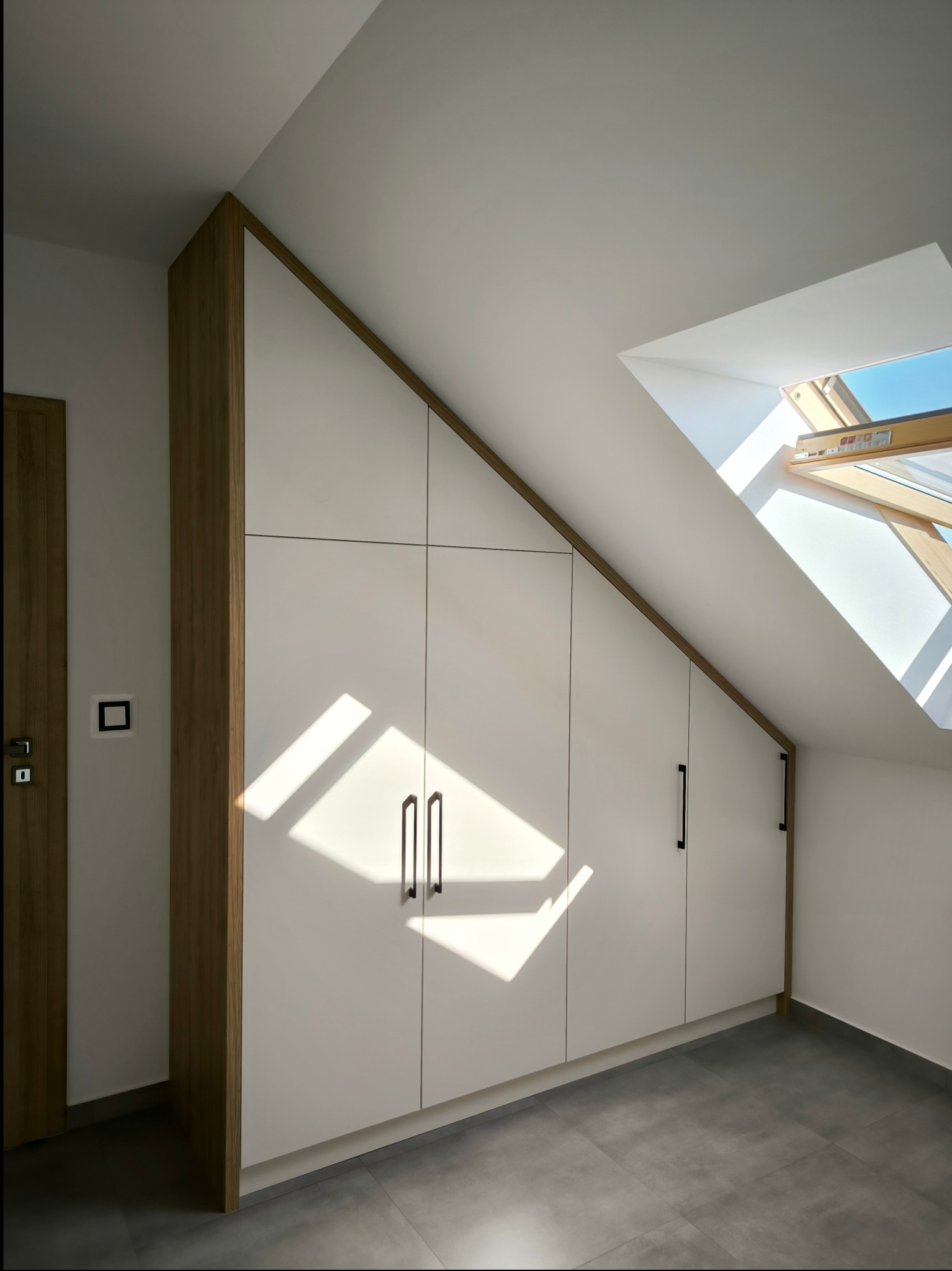 Biała szafa na wymiar z drewnianymi bokami, dopasowana do skosu poddasza, oświetlona promieniami słonecznymi wpadającymi przez okno dachowe.