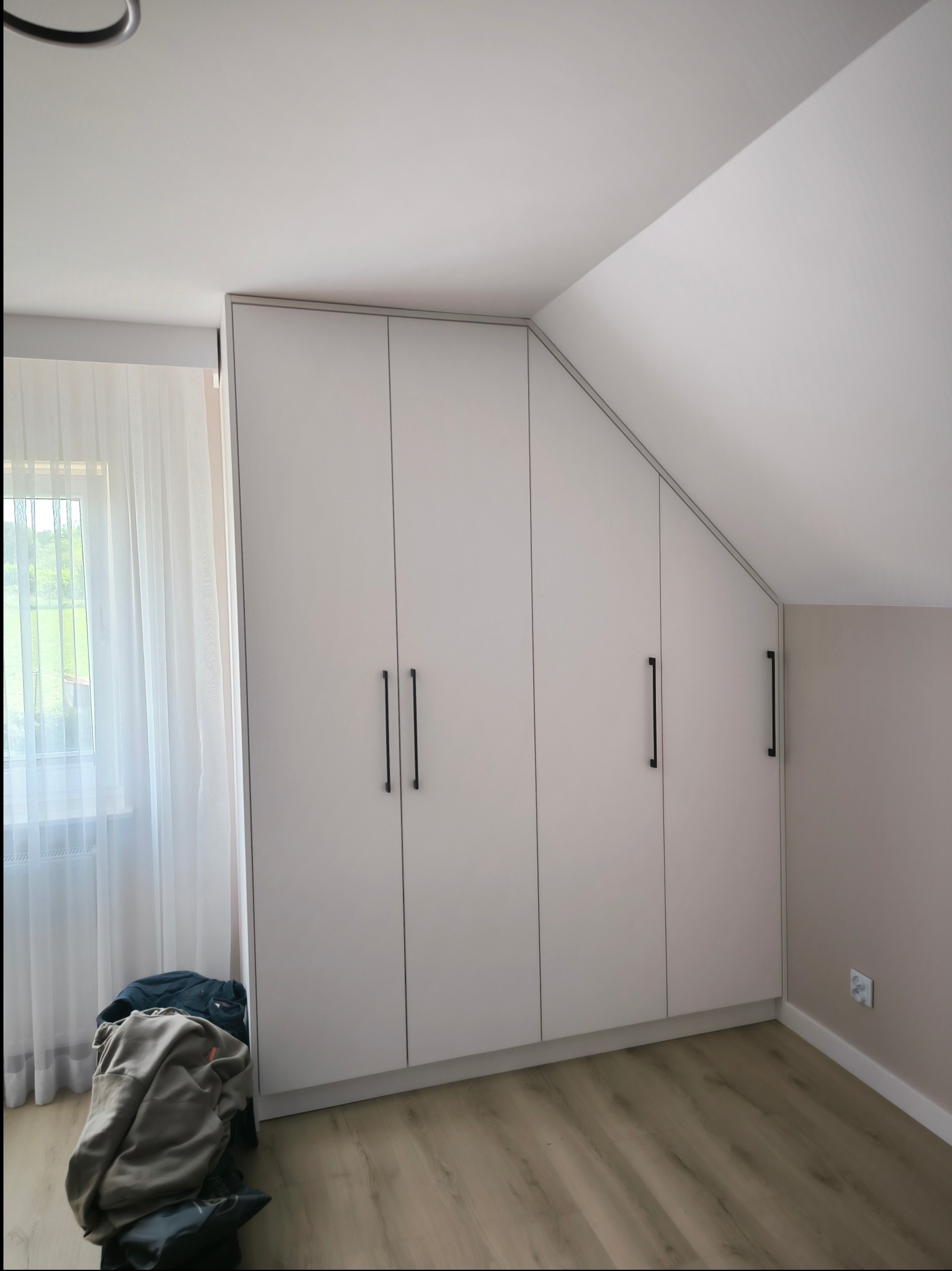 Biała szafa wnękowa na poddaszu z czarnymi uchwytami, widok na okno z zasłoną i stertę ubrań na podłodze