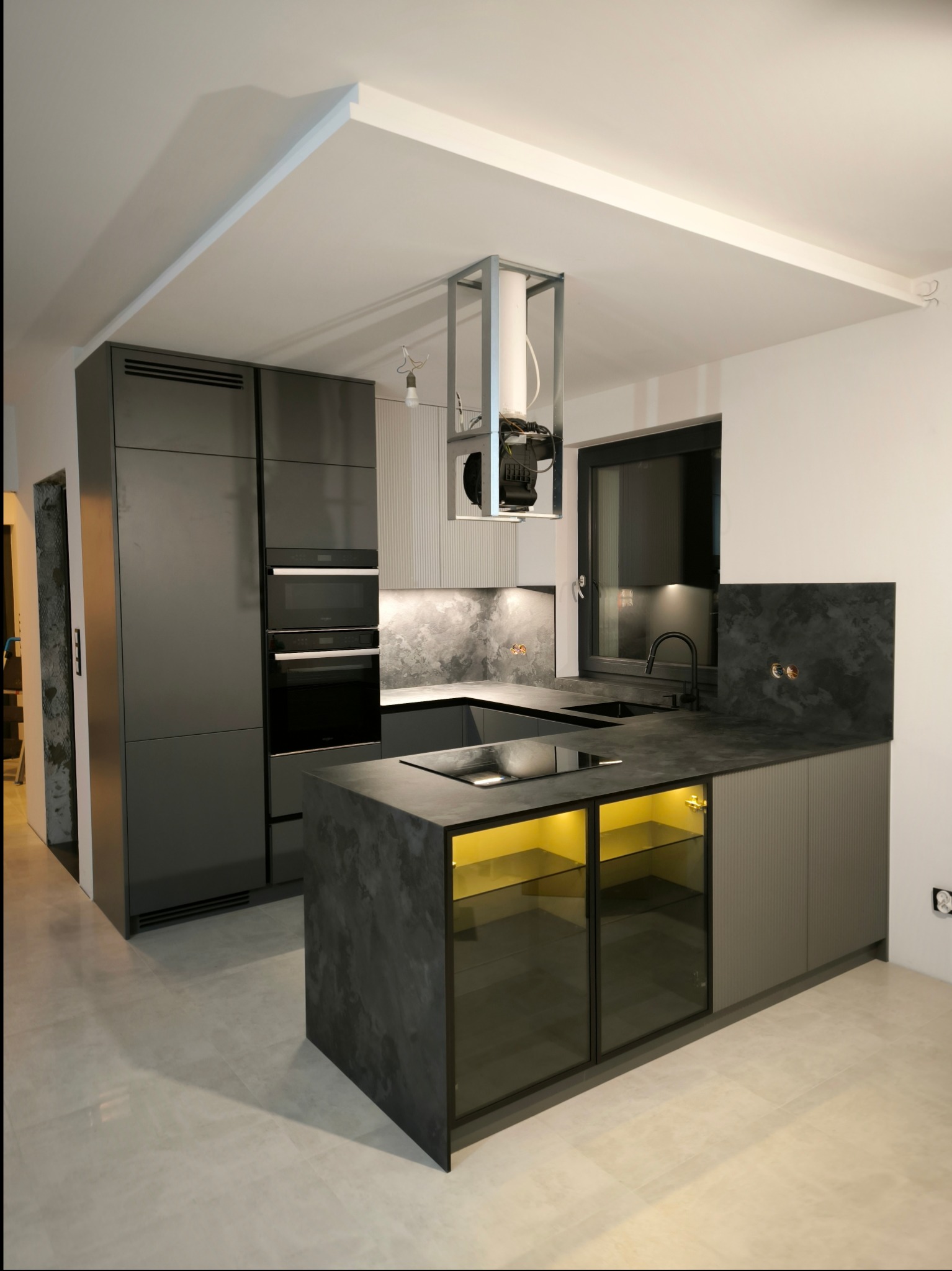 Nowoczesna kuchnia z szarymi frontami, wyspą z podświetleniem LED i czarnym blatem, widok na zabudowę piekarnika i okap z metalową konstrukcją.