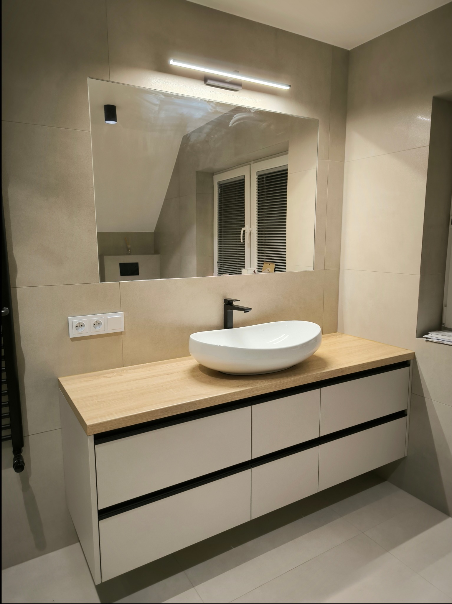 Nowoczesna łazienka z szafką pod umywalkę z jasnego drewna i białymi frontami z czarnymi akcentami, owalna umywalka nablatowa, lustro z podświetleniem LED, szare płytki na ścianach i podłodze.