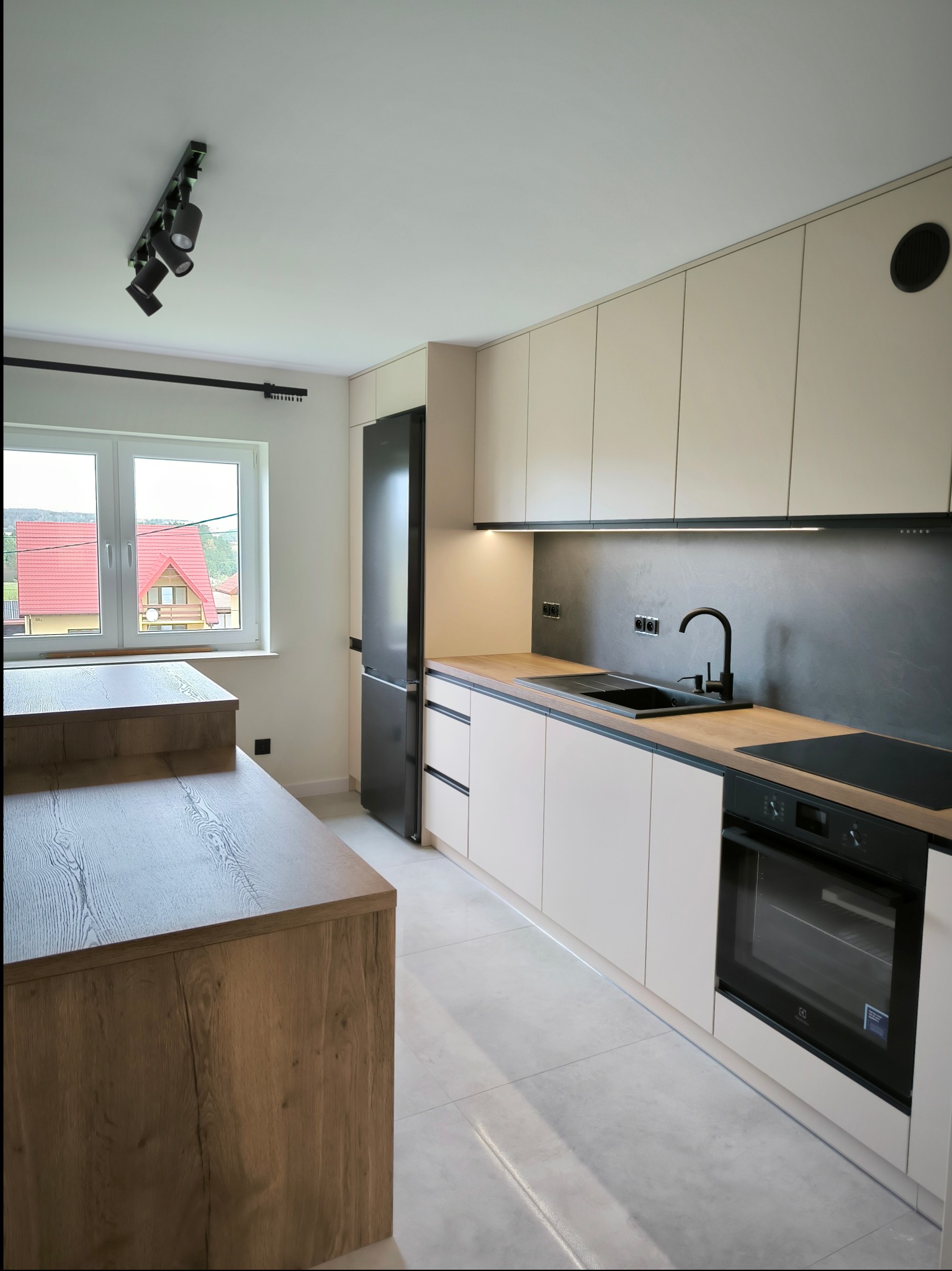 Nowoczesna kuchnia z beżowymi szafkami, czarnym zlewem i baterią, drewnianym blatem oraz wyspą kuchenną z widokiem na dom z czerwonym dachem za oknem.