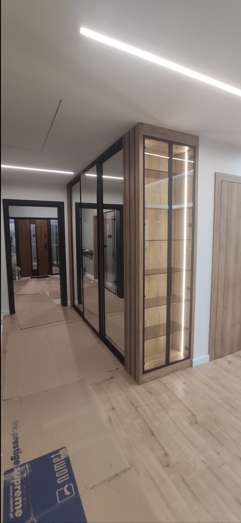 Nowoczesna szafa wnękowa z lustrzanymi drzwiami przesuwnymi i przeszklona witryna z oświetleniem LED, zabezpieczona kartonem na podłodze podczas montażu.