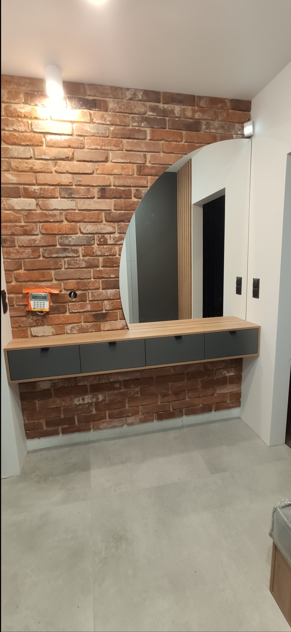 Loftowa aranżacja przedpokoju z szafką z szufladami w kolorze grafitowym, blatem w kolorze drewna i okrągłym lustrem na ceglanej ścianie.