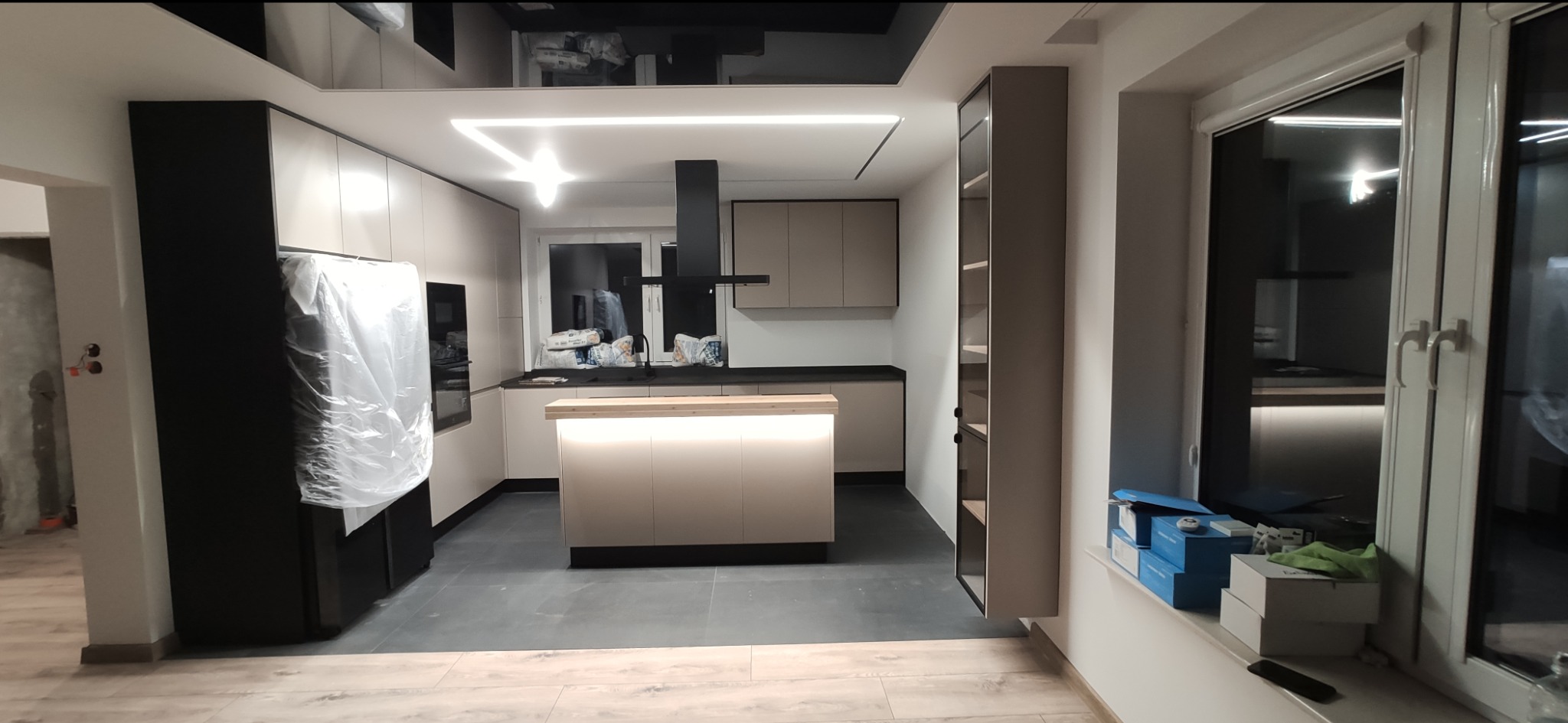 Nowoczesna kuchnia w trakcie montażu: wyspa z podświetleniem LED, czarny okap, zabudowa w odcieniach beżu i czerni, lodówka zabezpieczona folią, widoczne materiały budowlane przy oknie.