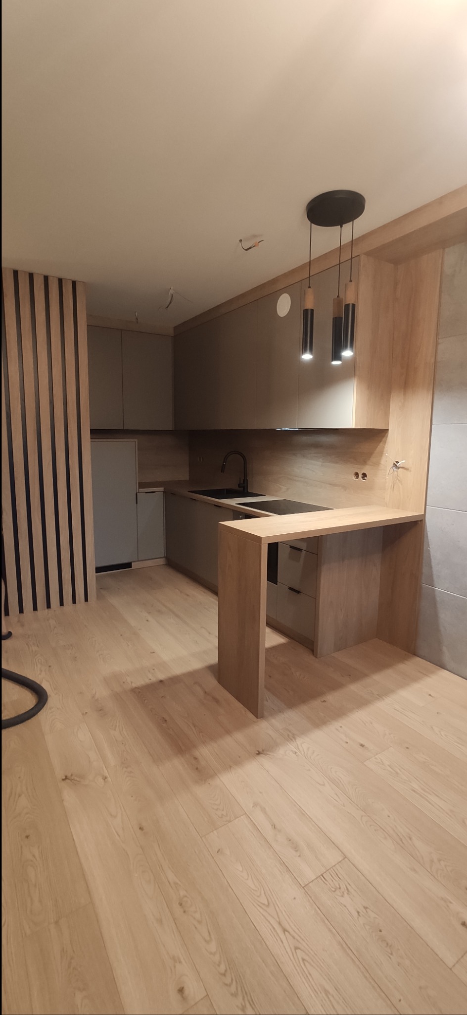 Nowoczesna kuchnia z drewnianymi akcentami, szare fronty szafek, drewniany blat z barkiem, podwieszane lampy i dekoracyjne drewniane panele pionowe na ścianie.