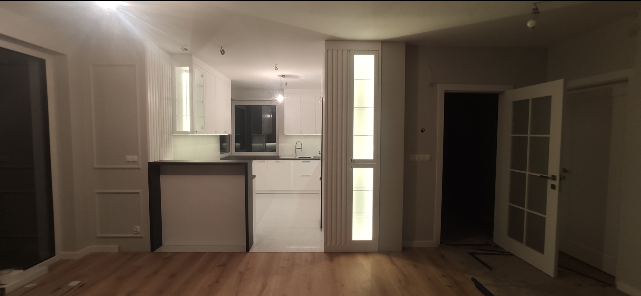 Nowoczesna, biała kuchnia z wyspą i oświetleniem LED, widok z salonu na otwartą przestrzeń, drewniana podłoga, białe ściany z dekoracyjnymi panelami.