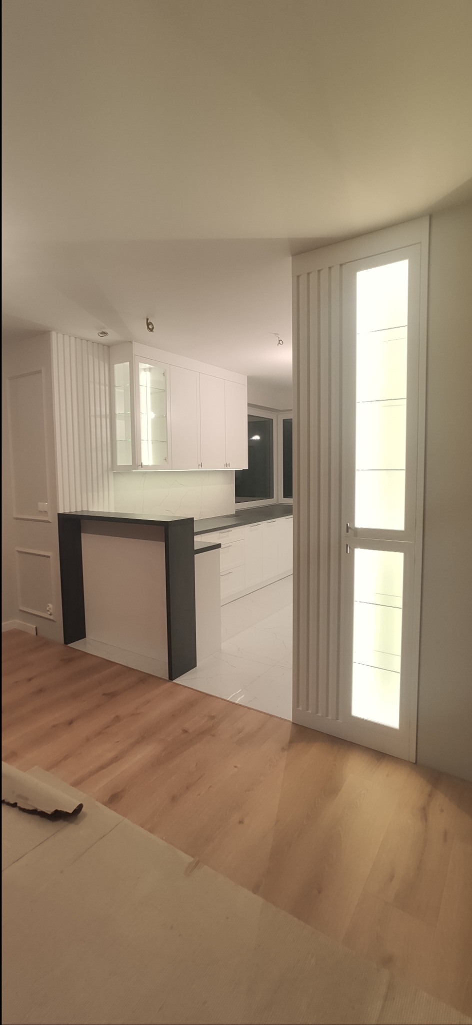 Nowoczesna, biała kuchnia z czarnym blatem oddzielona od salonu barkiem, widok z perspektywy podłogi z drewnianych paneli.