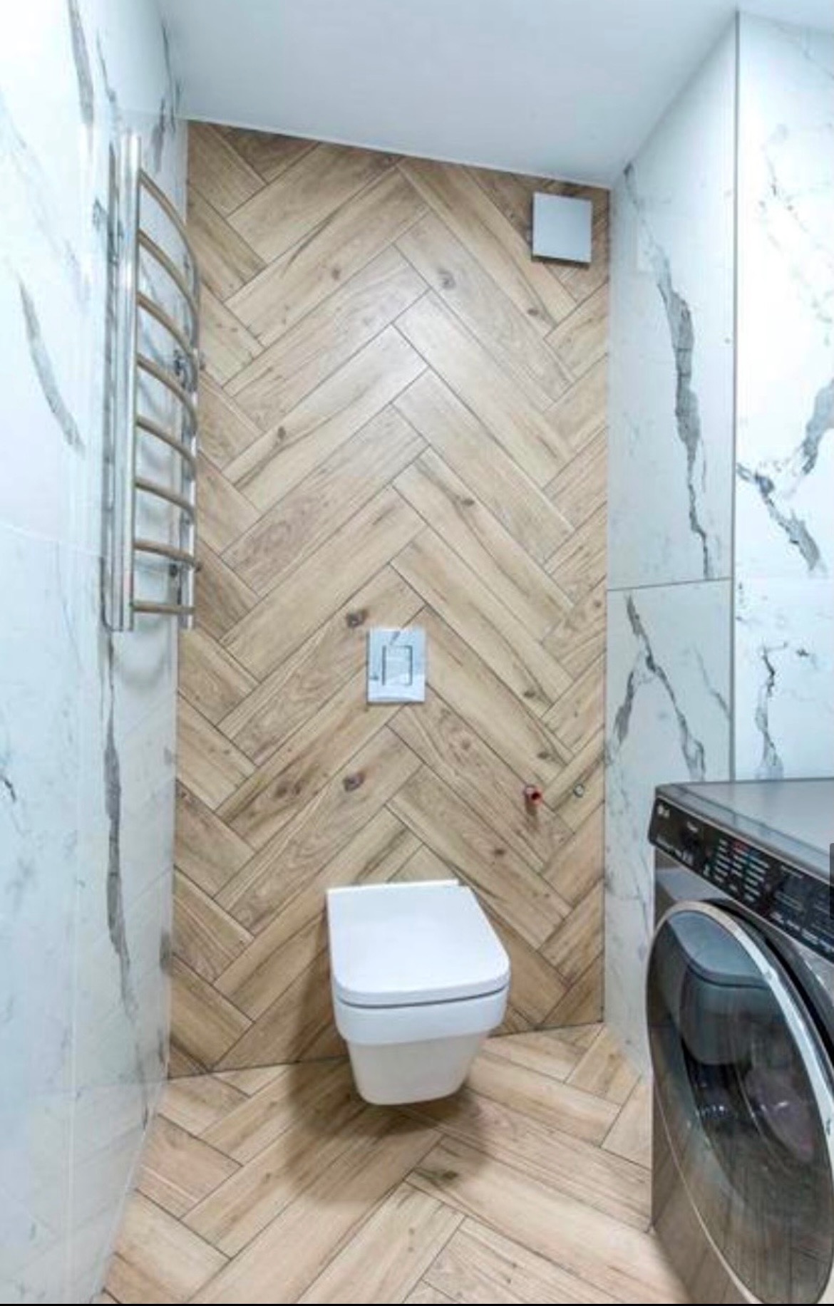 Nowoczesna łazienka z drewnopodobną glazurą ułożoną w jodełkę na ścianie i podłodze, wisząca toaleta, marmurowe płytki na ścianach, grzejnik drabinkowy i pralka.