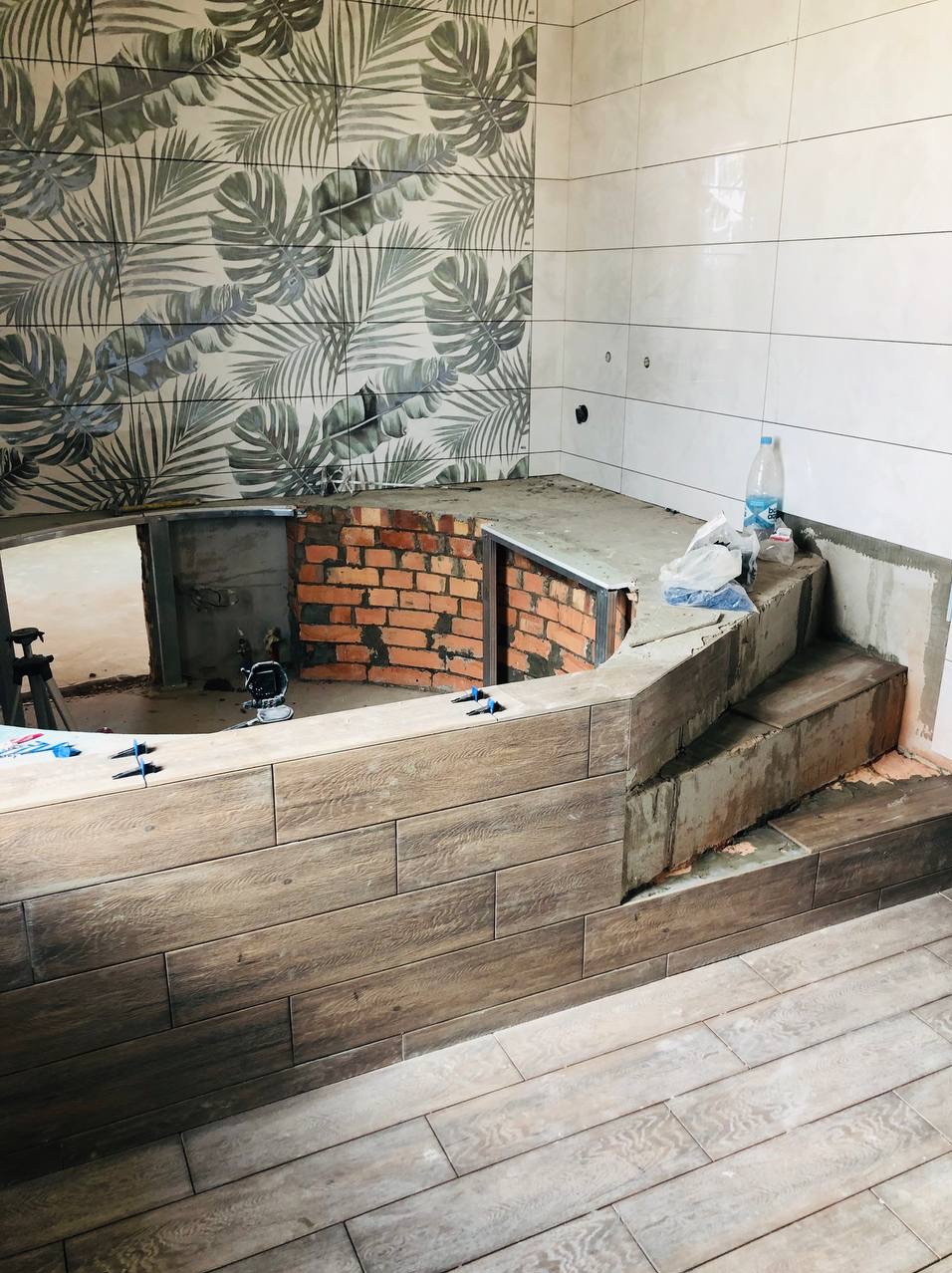 Wnętrze łazienki w trakcie remontu: obudowa wanny z brązowych płytek imitujących drewno, ściana z białych płytek i płytek z motywem liści palmowych, widoczne narzędzia i materiały budowlane.