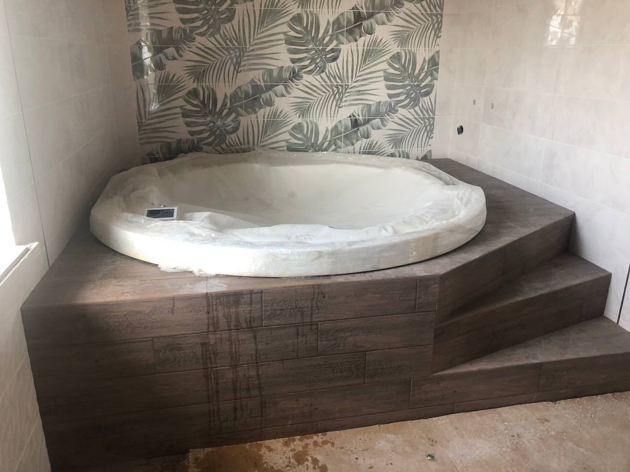 Okrągła wanna w folii ochronnej, obudowana płytkami imitującymi drewno, ściany wyłożone białymi płytkami i płytkami z motywem liści tropikalnych, widoczne fragmenty podłogi przed wykończeniem.