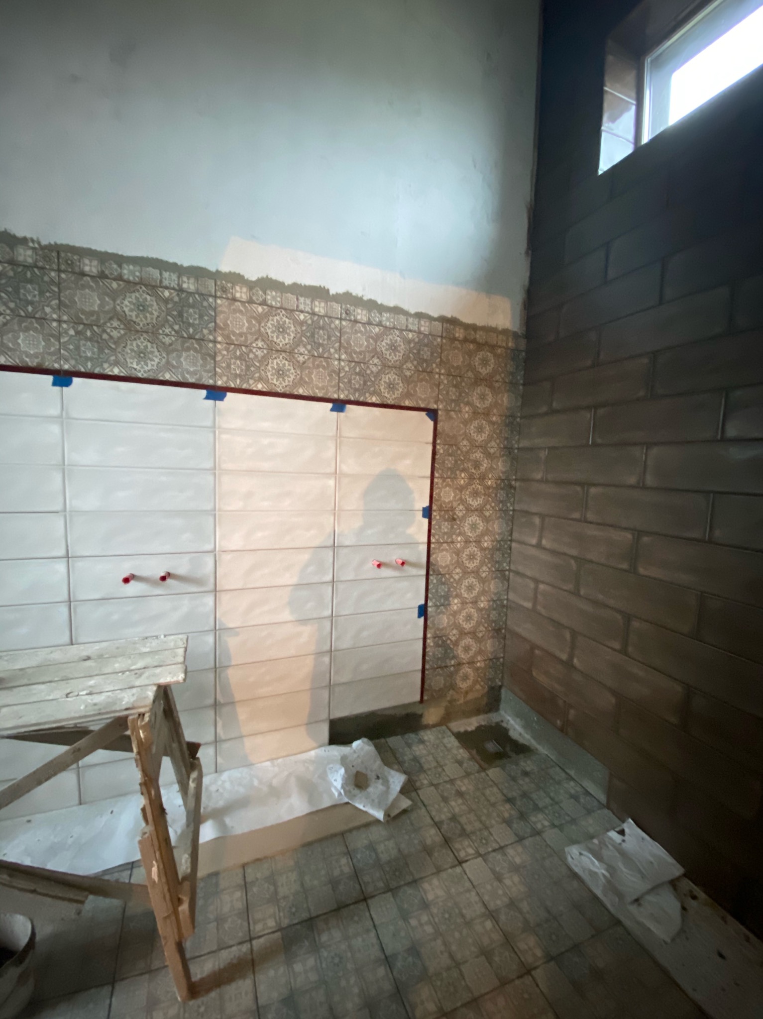 Ściana łazienki w trakcie układania płytek: częściowo ułożone białe płytki z krzyżykami dystansowymi i niebieską taśmą, powyżej dekoracyjne płytki z wzorem, obok ściana wyłożona ciemnymi płytkami...