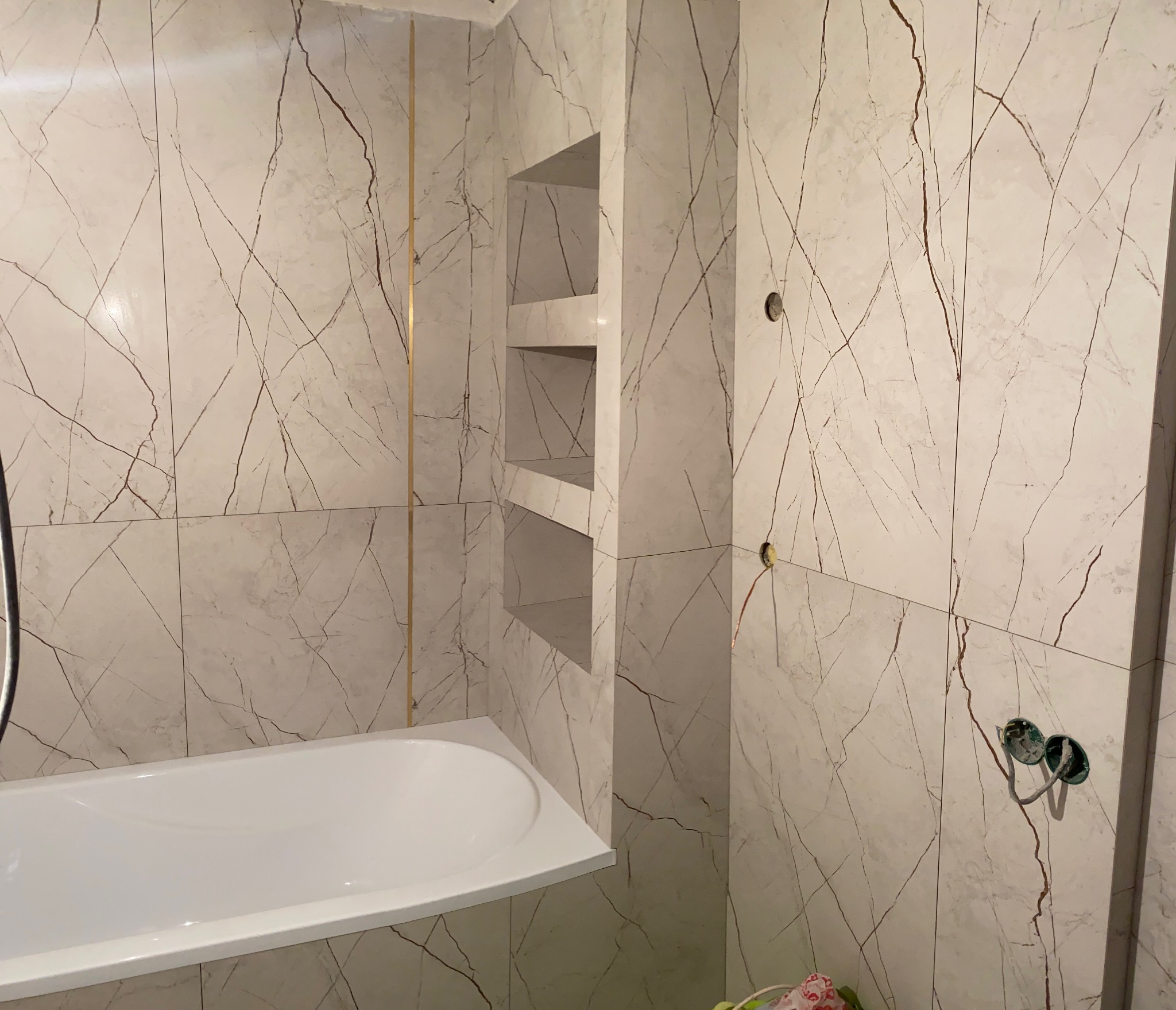 Biała wanna z obudową, ściany wyłożone płytkami imitującymi marmur z żyłkami w kolorze brązowym, wnęka z półkami wykończona tymi samymi płytkami, wystające kable elektryczne w ścianie.