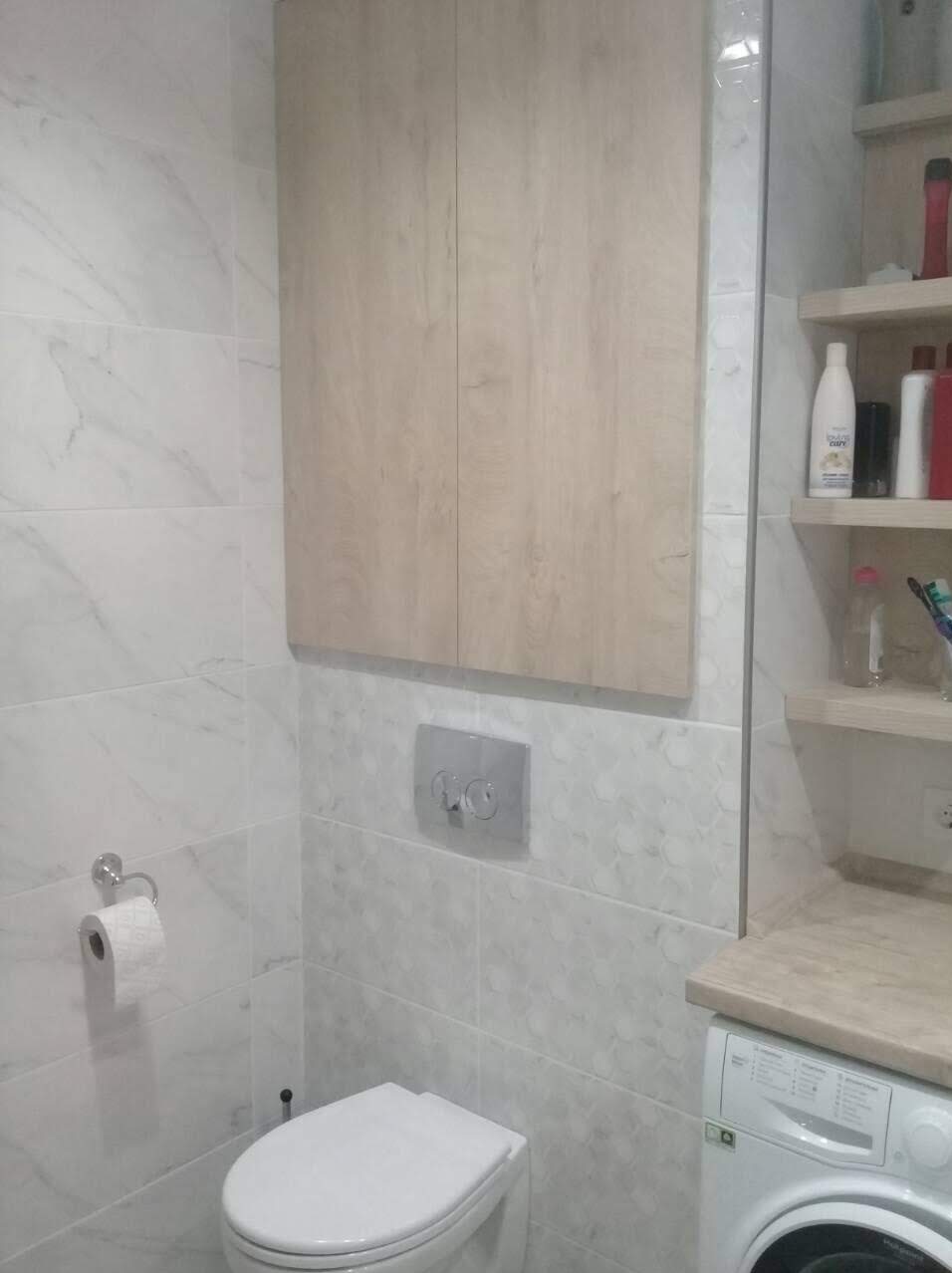 Jasna łazienka z marmurkowymi płytkami, wisząca szafka z drewnianymi frontami nad toaletą, chromowany przycisk spłuczki i półki z kosmetykami po prawej stronie.