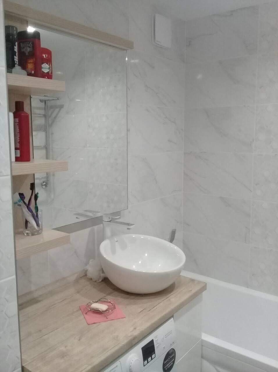 Jasna łazienka z marmurowymi płytkami, okrągła umywalka nablatowa, drewniany blat i pralka pod blatem; na półkach kosmetyki i szczoteczki do zębów.