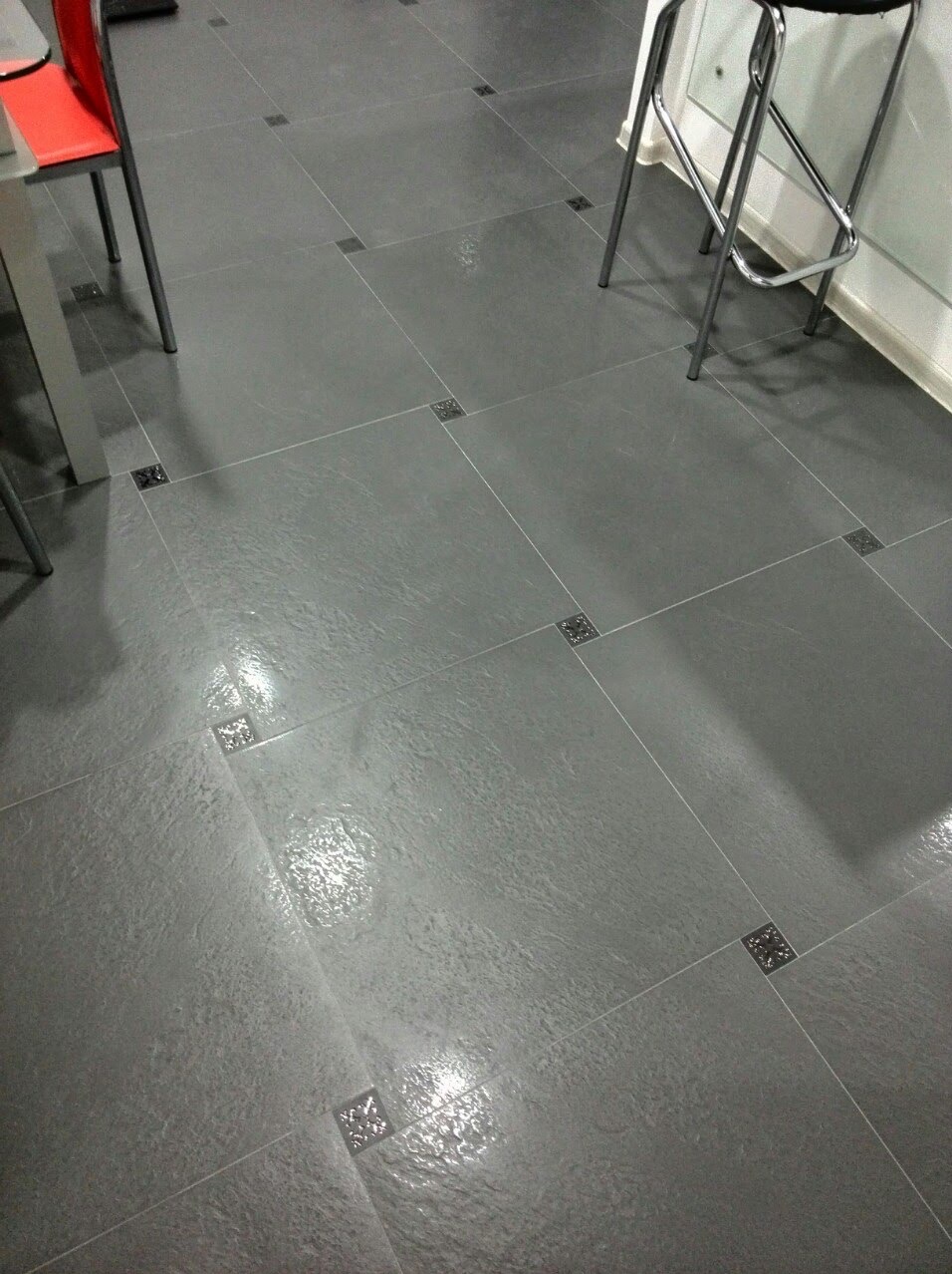 Szare płytki podłogowe z dekoracyjnymi wstawkami w spoinach, widoczne fragmenty krzesła i stołka barowego.