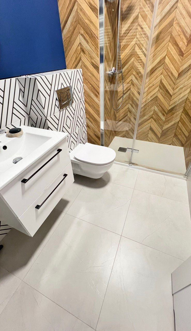 Nowoczesna łazienka z drewnianą mozaiką na ścianie prysznica, białą ceramiką i umywalką z czarnymi uchwytami, akcentowana niebieską ścianą.