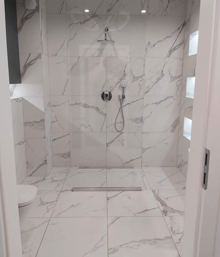 Kabina prysznicowa walk-in wykończona białymi płytkami imitującymi marmur, z odpływem liniowym i wnękami z podświetleniem LED.